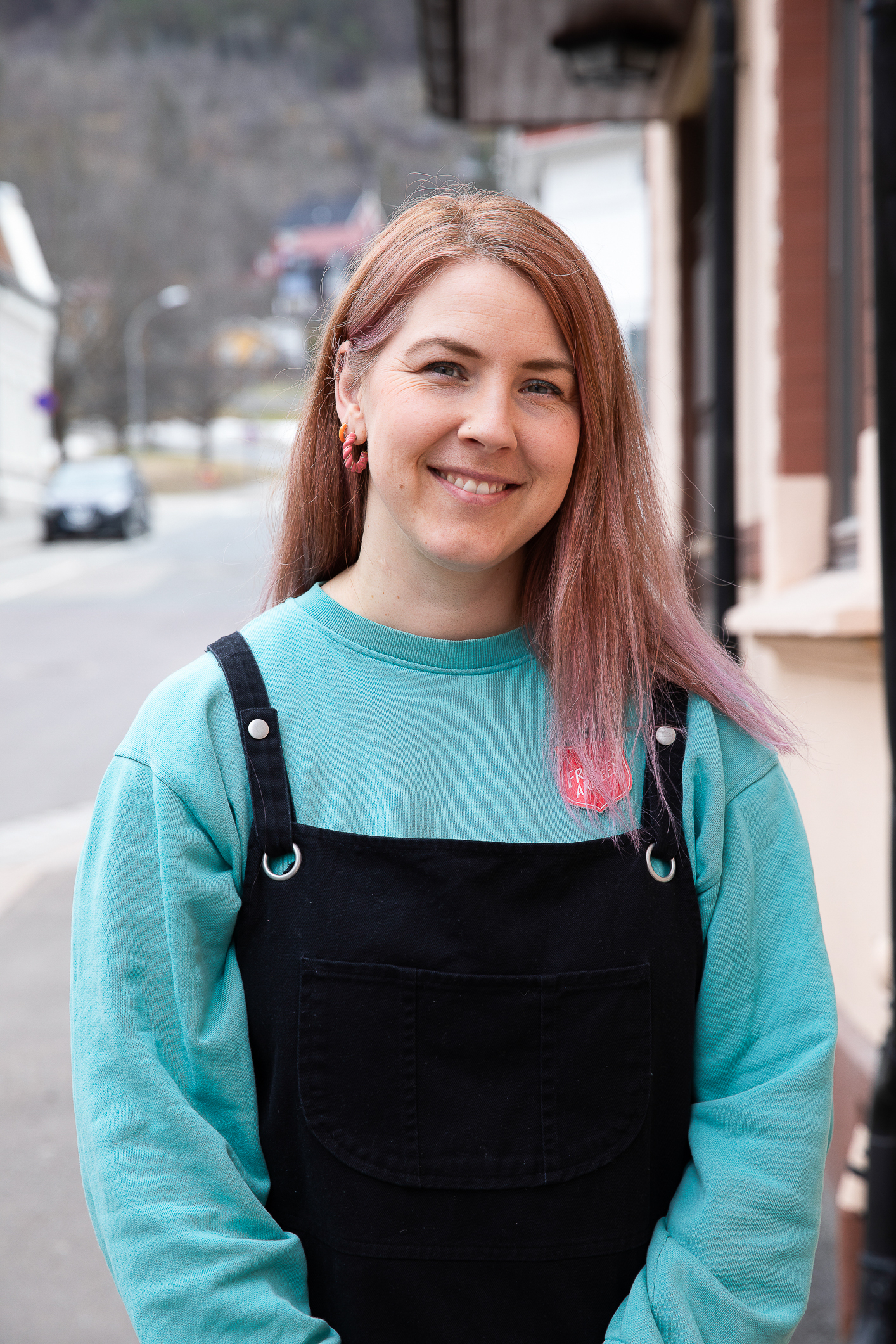 Foto: Kvinne tidlig i 40-årene med turkis genser og svart selebukse står ute i bybildet.Hun har langt hår med litt rosa skjær i og smiler mot kamera.