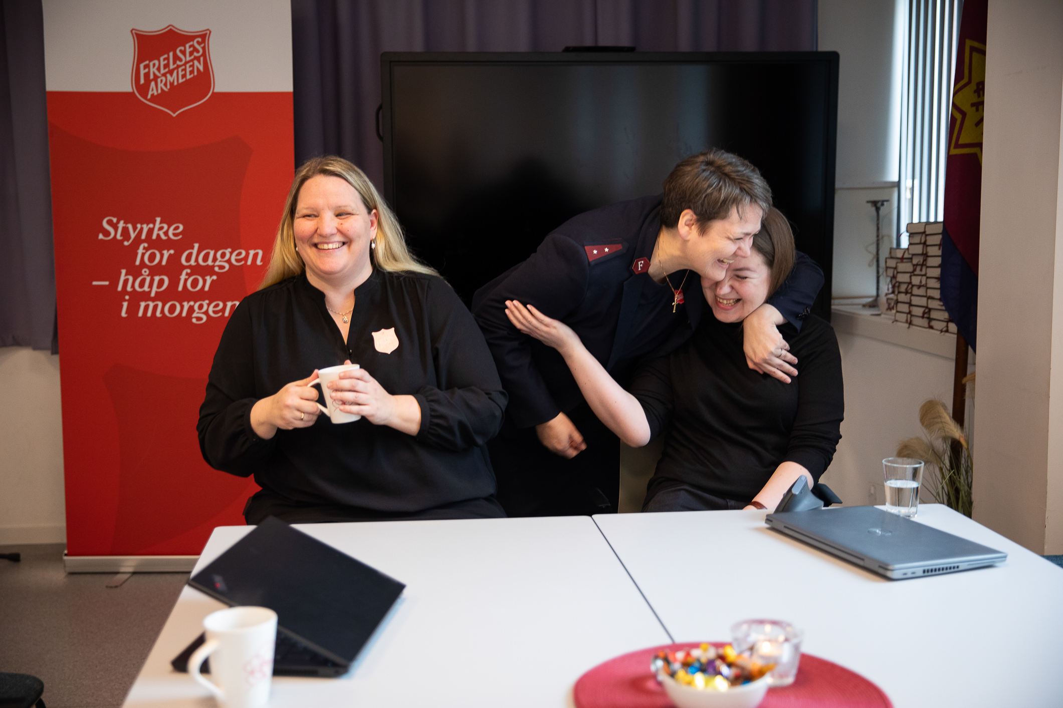 Foto: To kvinner sitter bak et bord i et kontorlokale. Mellom dem står en kvinne i uniform fra Frelsesarmeen. som bøyer seg ned og gir kvinnen til høyre en klem. Den andre kvinnen kommer også fra Frelsesarmeen og holder en hvit kopp i hånden.