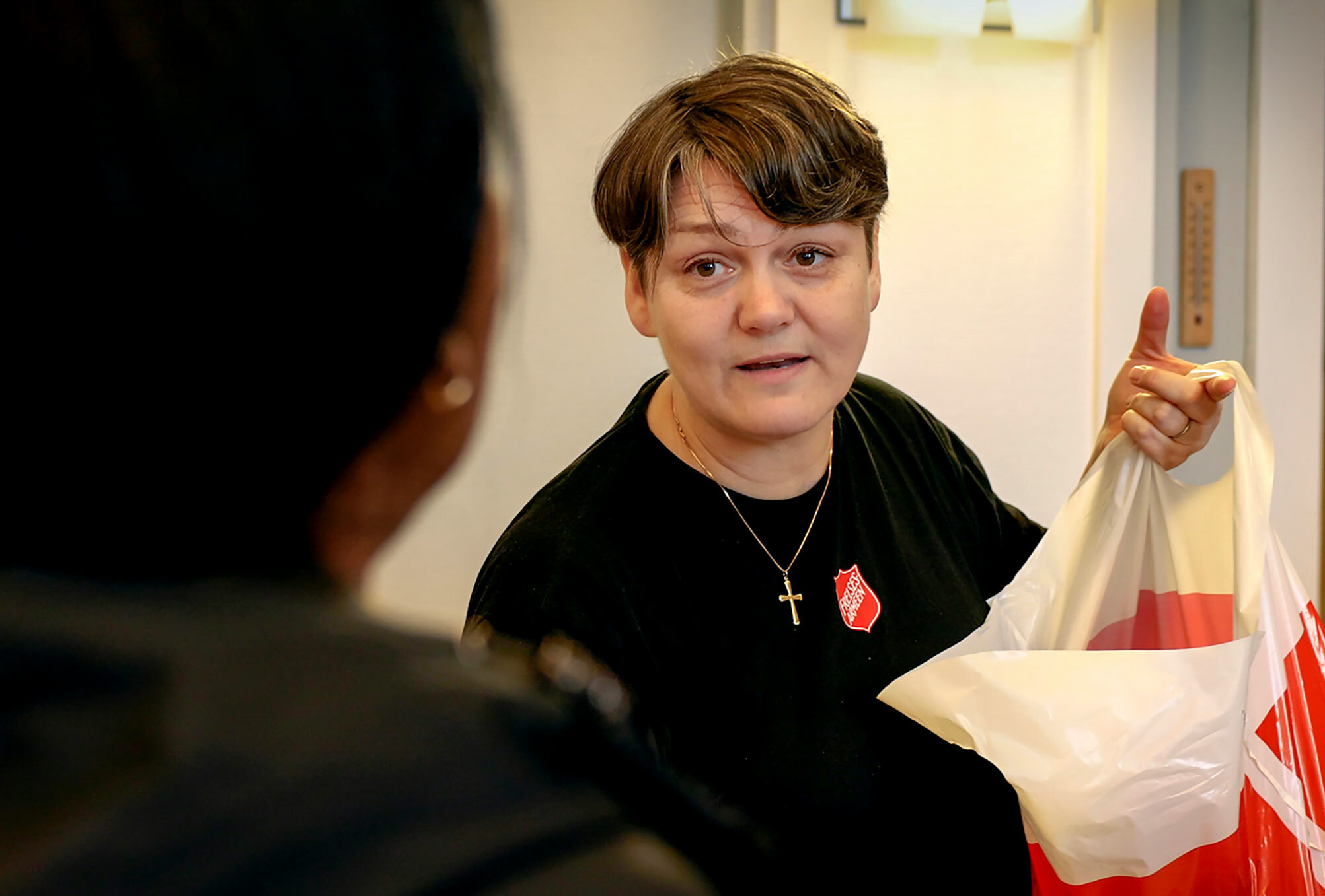 Foto: Nærbilde av ansikt til kvinne som jobber i Frelsesarmeen. Hun har på seg mørk genser med rød logo og et kors rundt halsen. Ute i høyre hjørne sees litt av en rød og hvit plastpose.