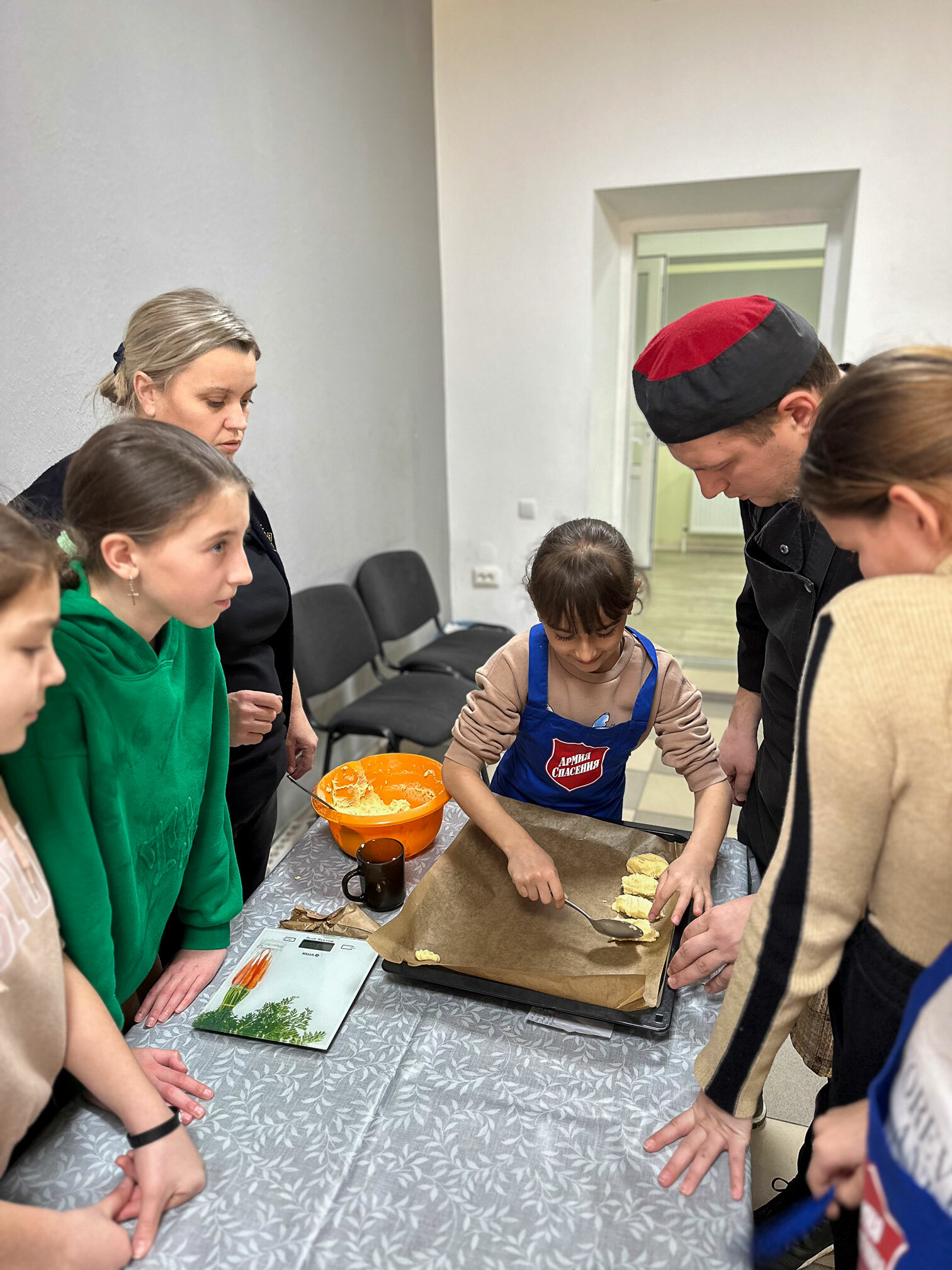 Foto: En jente legger ut klater med kakedeig på et stekebrett. Rundt bordet står et par andre voksne og flere barn.