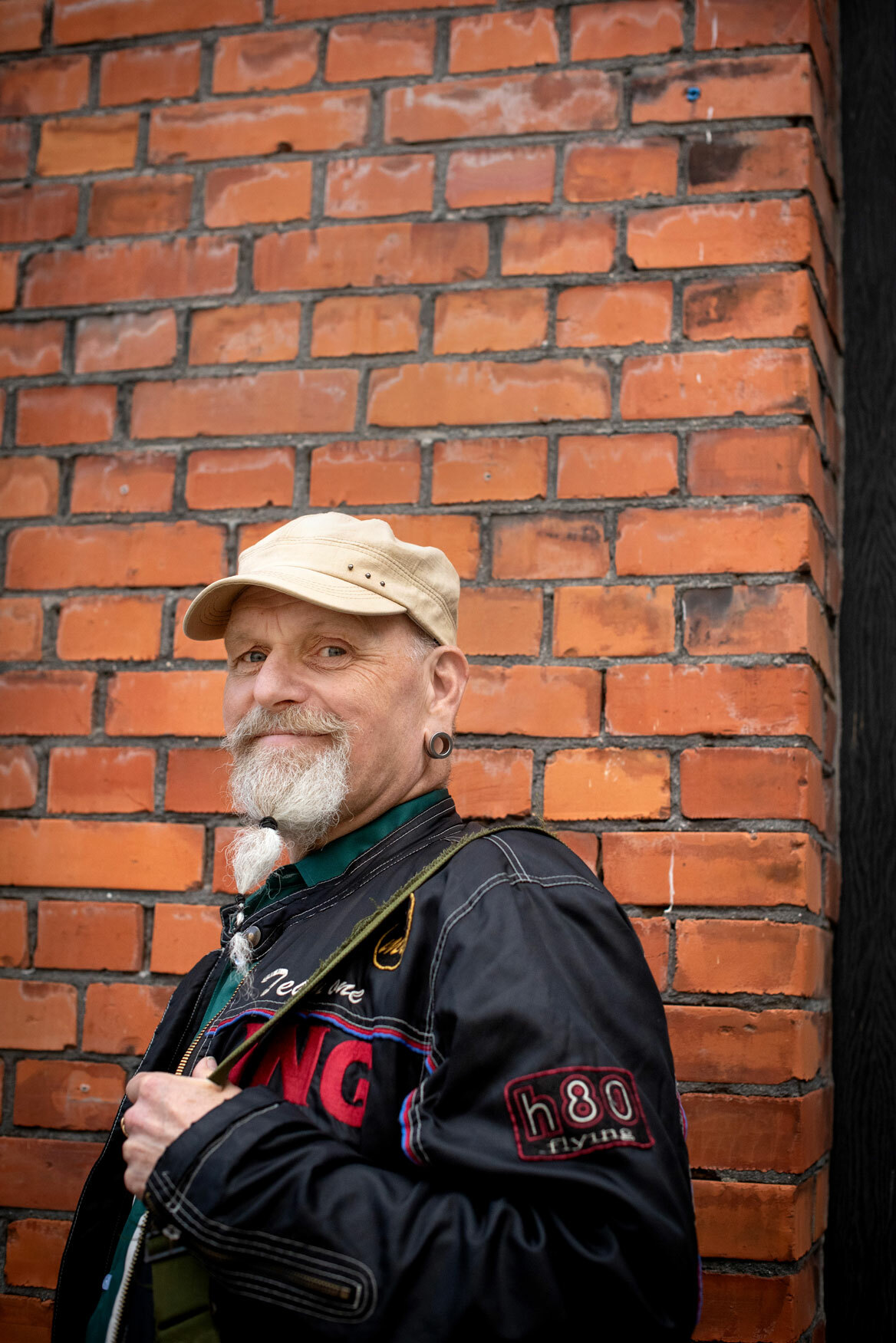 Foto: Mann i skinnjakke, caps og langt, grått skjegg står lent opp mot en rød musteinsvegg.