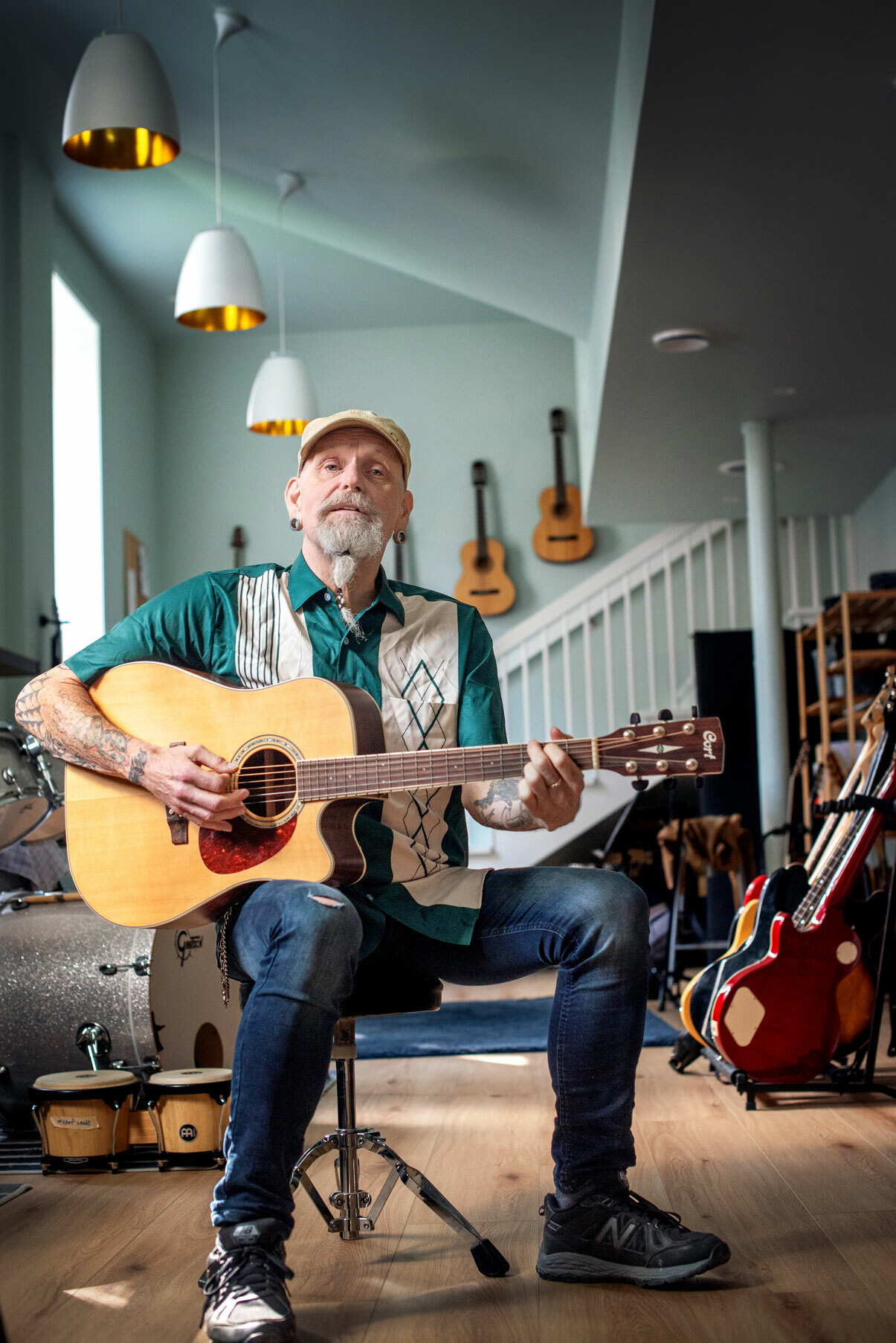 Foto: Mannen med skjedd og caps spiller gitar. Han sitter i et rom fylt med instrumenter.