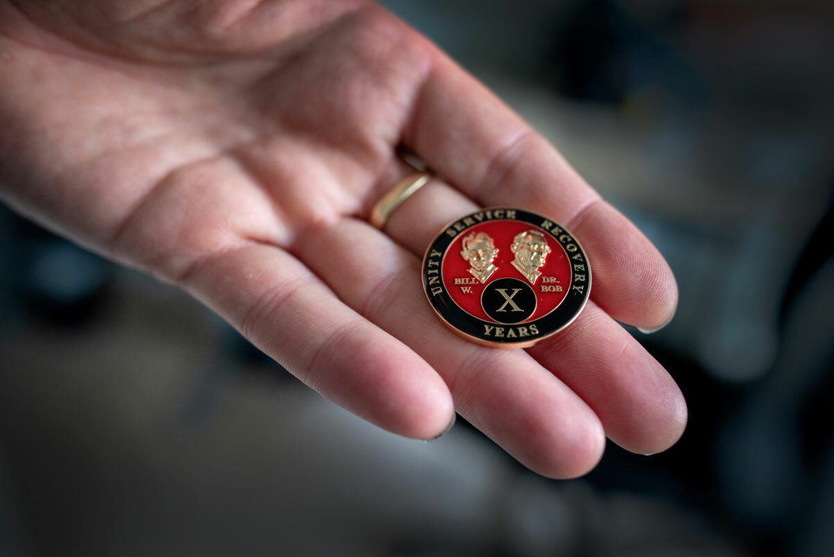 Foto: Mannshånd holder fram en pin i svart, rødt og gull med en X foran.