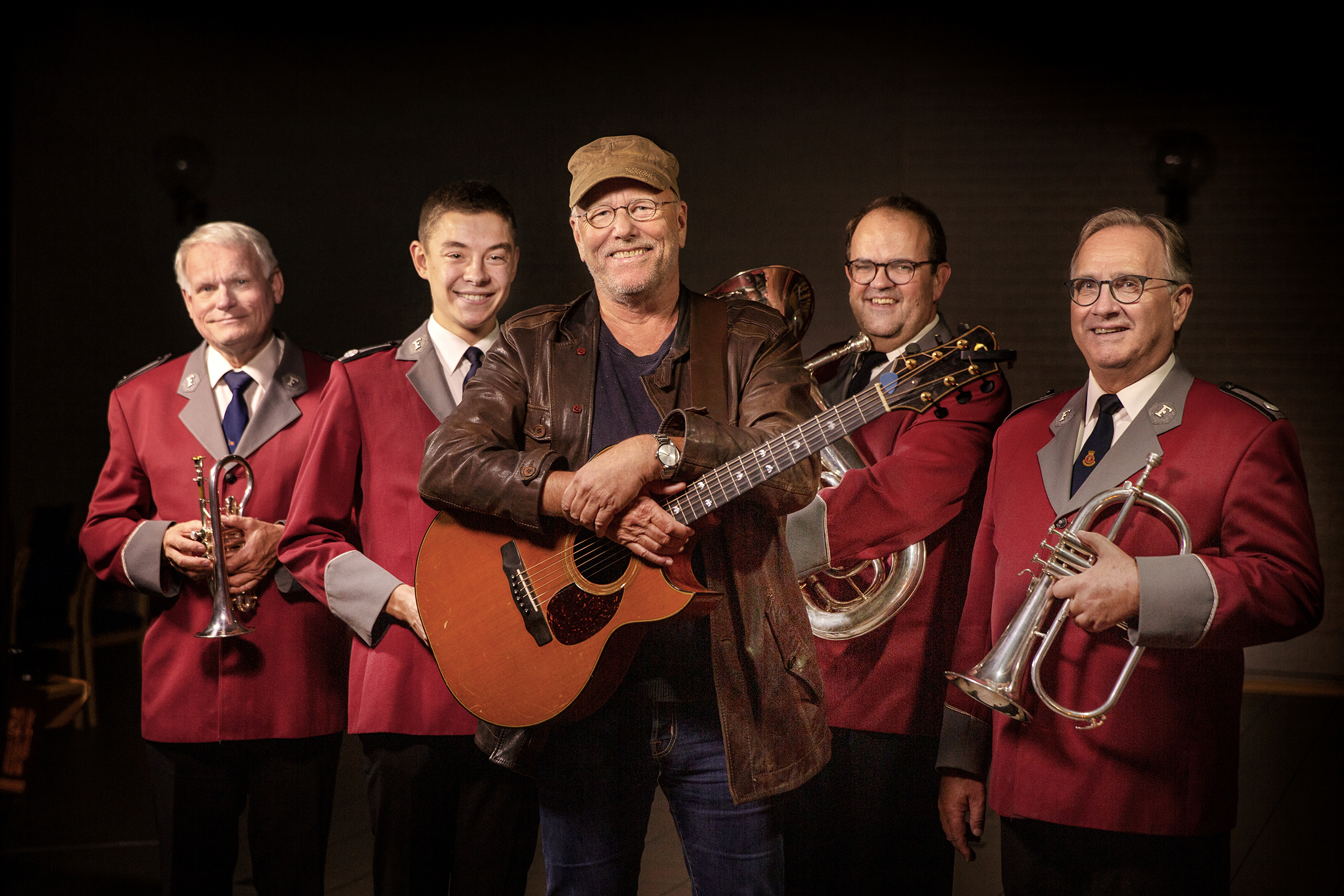 Bildet viser Lars Martin Myhre som holder gitaren sin. Bak ham står fire musikere fra Frelsesarmeen i røde jakker og holder forskjellige instrumenter.