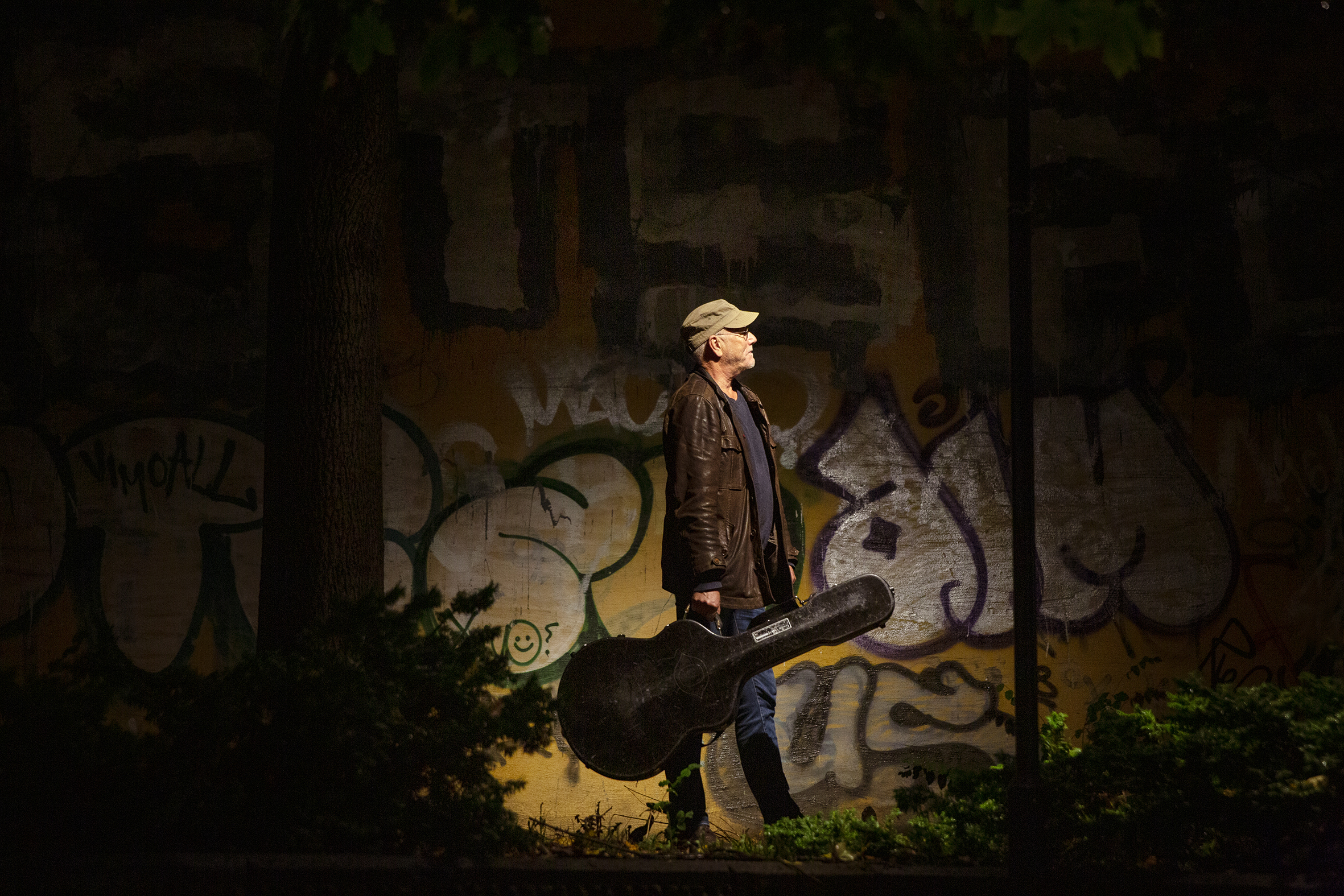 Bildet er tatt litt på avstand og viser Myhre forn en vegg med grafitti. Han står med siden til kamera, og holder en gitarkasse.