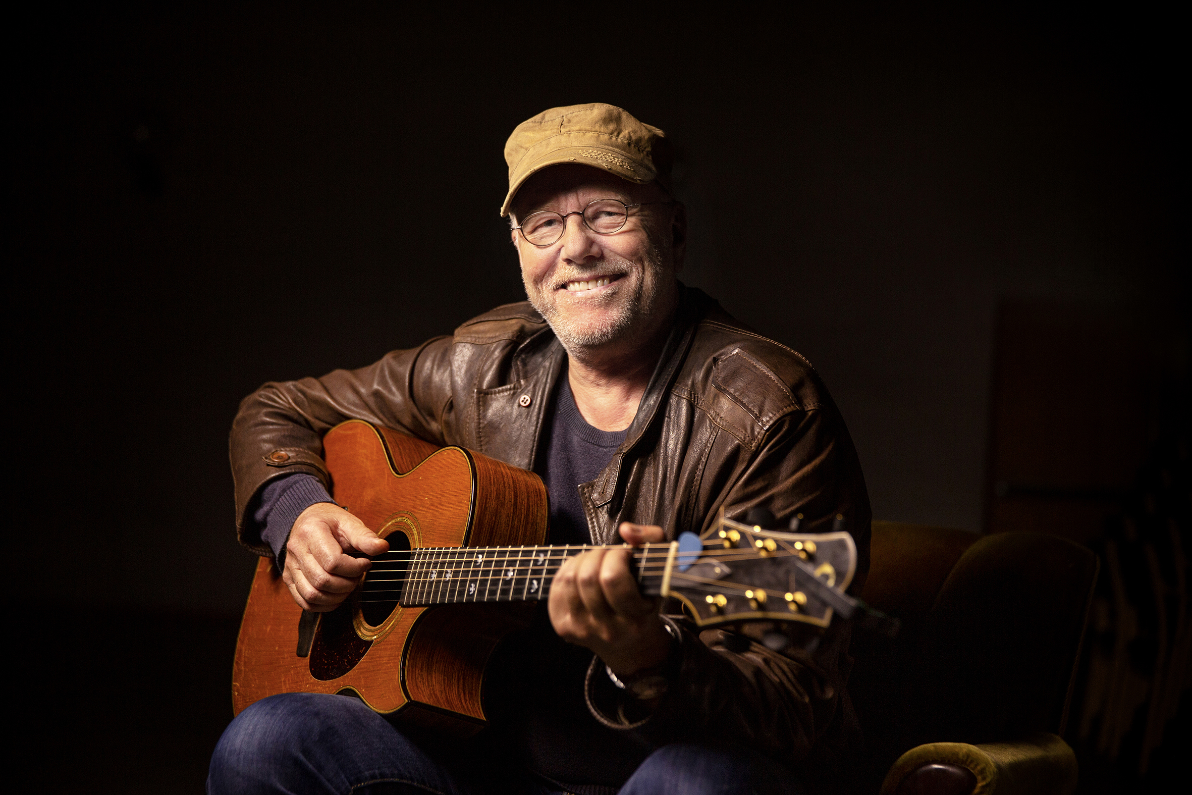 Bildet viser Lars Martin Myhre som sitter og spiller gitar, han har på seg caps og brun skinnjakke.