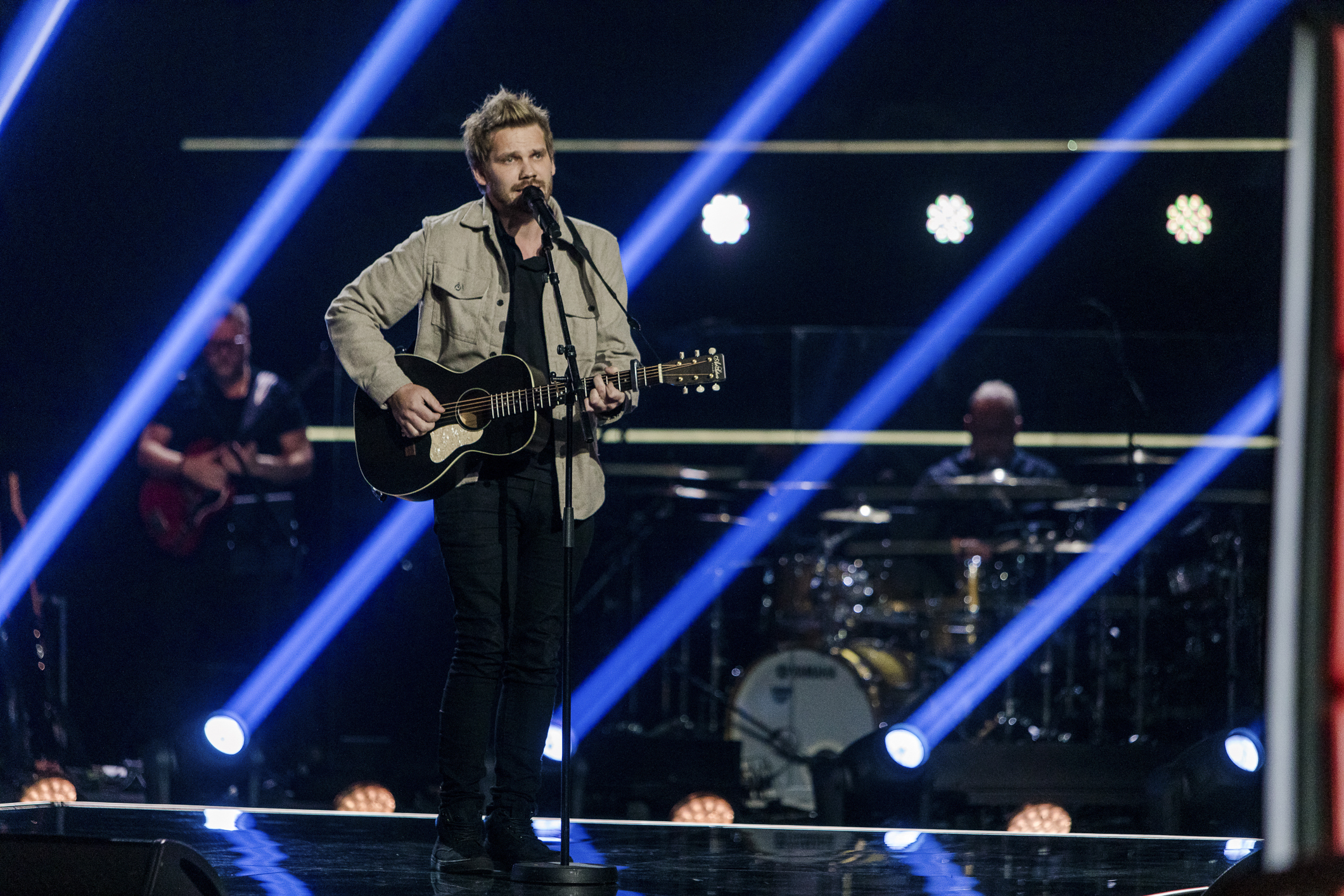 En ung mann med gitar og mikrofon står på en scene med blått lys bak. Han har på en kakifarget jakke og mørk bukse. Håret er kort og stylet. Han synger og spiller på gitar.