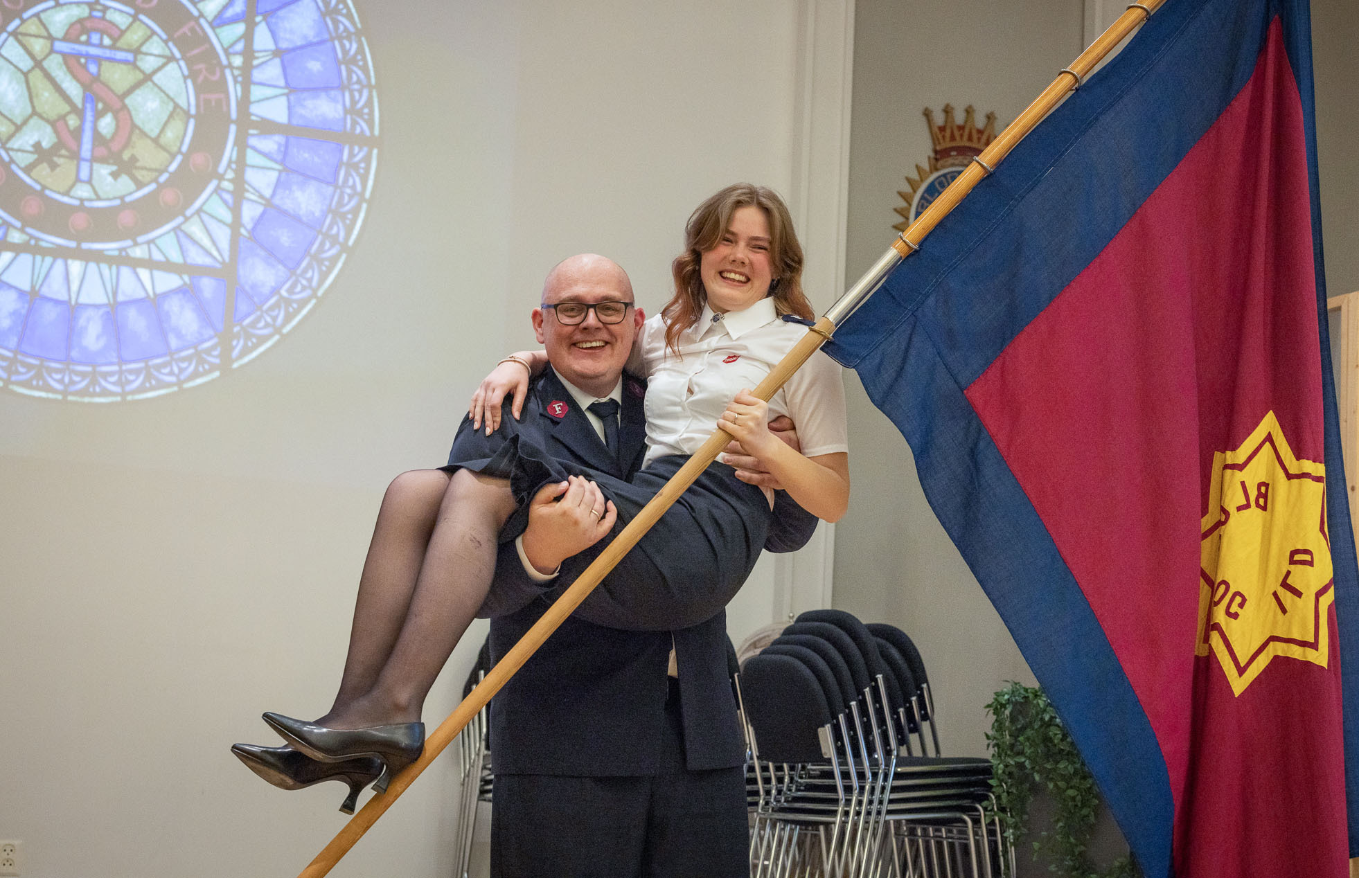 Far har løftet opp tenåringsdatter og holder henne i sine armer som et mindre barn. Begge er i uniform. I bakgrunnen ses et flagg fra Frelsesarmeen og et vindu med glasskunst.