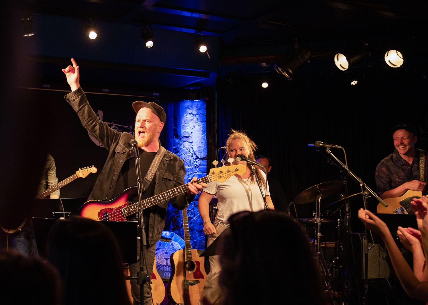 Vokalist som synger i mikrofonen. Han har en gitar i hendende. Bandet står i bakgrunnen i det mørke lokalet.