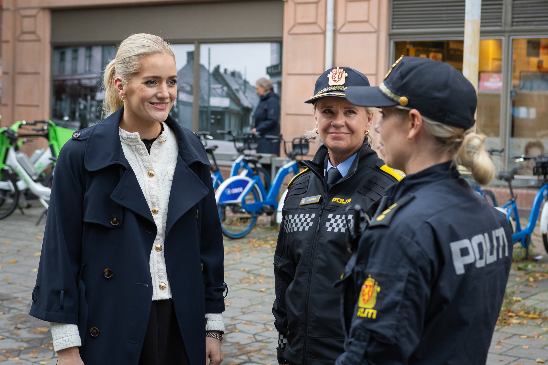 Tre kvinner snakker sammen og smiler. To i sort politiuniform med skyggeluer. Kvinnen til venstre har lyst hår og hestehale og blå kåpe med hvit genser inni.