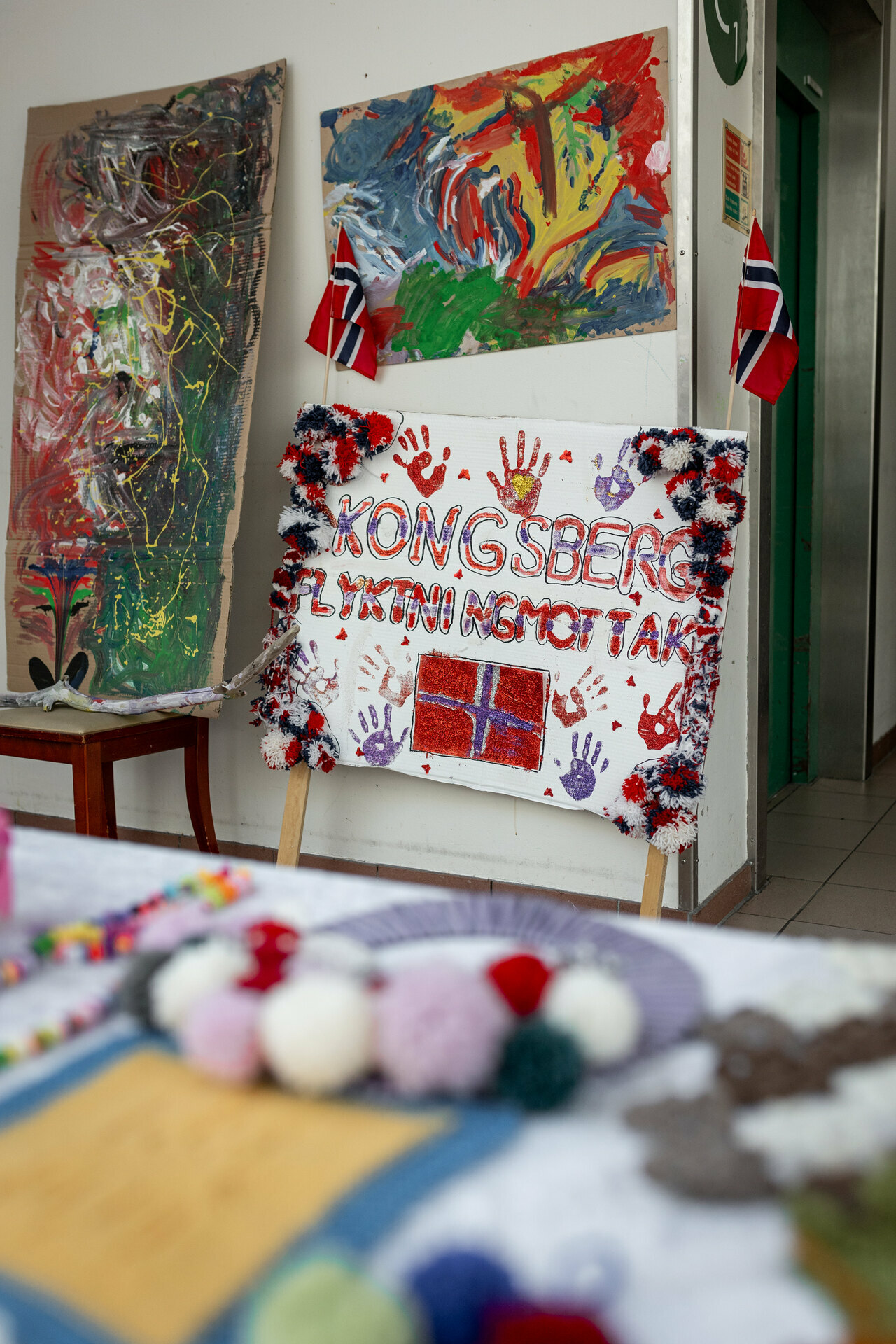 Foto: Av malerier, blant annet et skilt med det norske flagget der det står Kongsberg flyktningmottak over og barnehender.