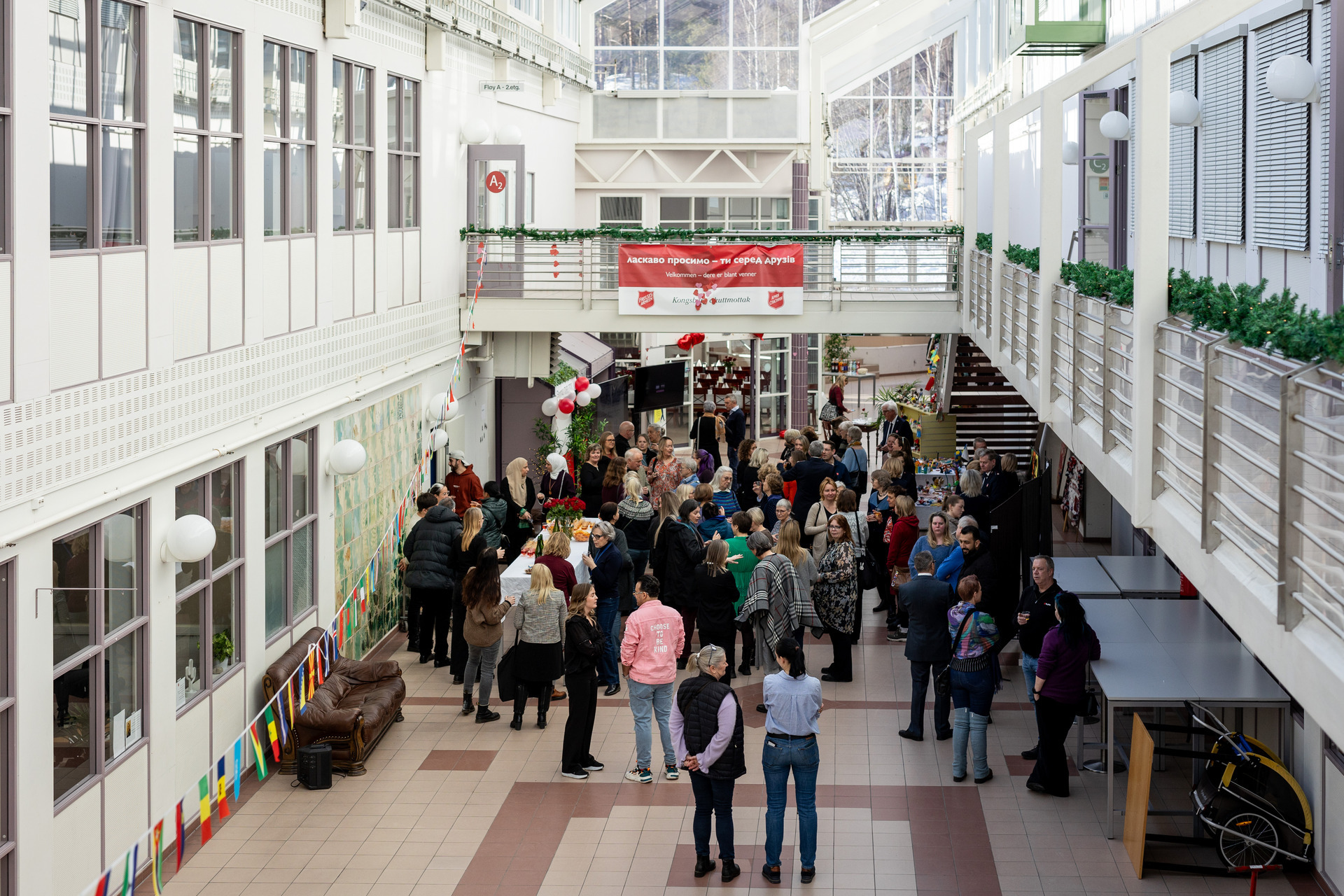 Foto: To bilder etter hverandre. Viser sammen store rom med glasstak høyt over – med og uten folk. På det første bildet er en lang rekke med flaggvimpler i fokus.