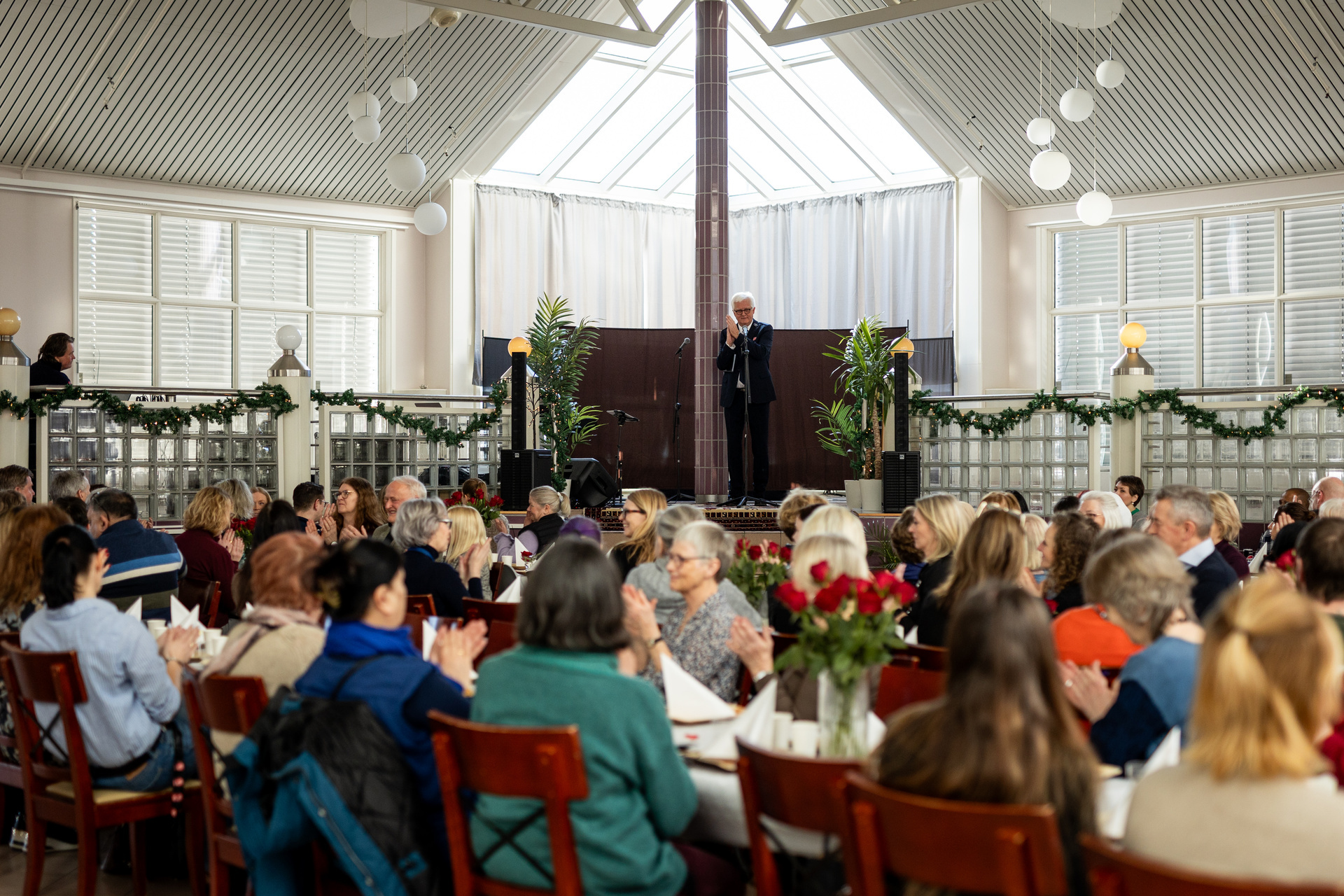Foto: Mann i uniform står på et podium og snakker i enden av lokalet. På bordene foran sitter mange mennesker.