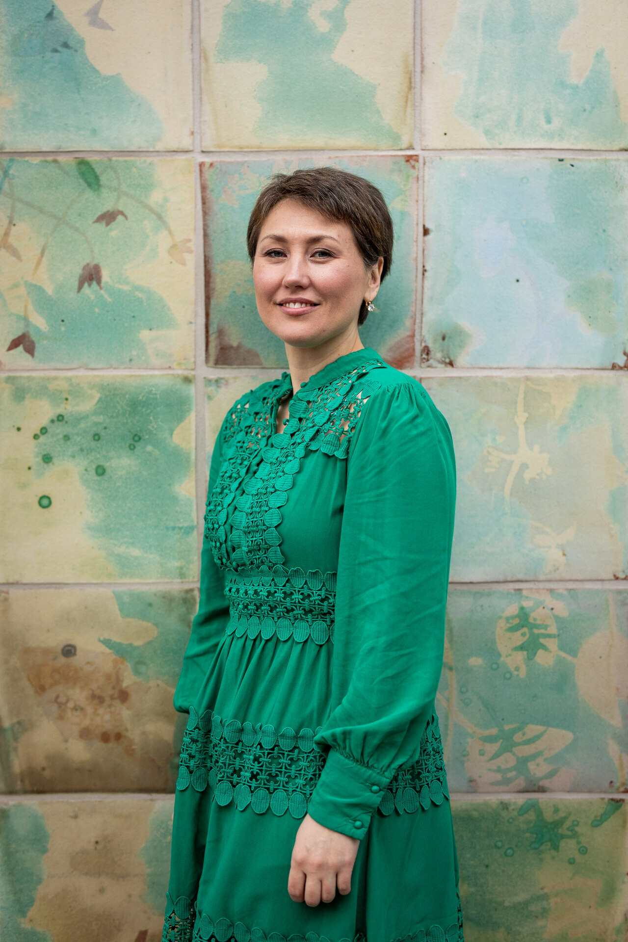 Foto: Av kvinne i 30-årene med kort, mørkt hår og grønn kjole. Hun smiler og står foran en vegg med sjøgrønne, store fliser.