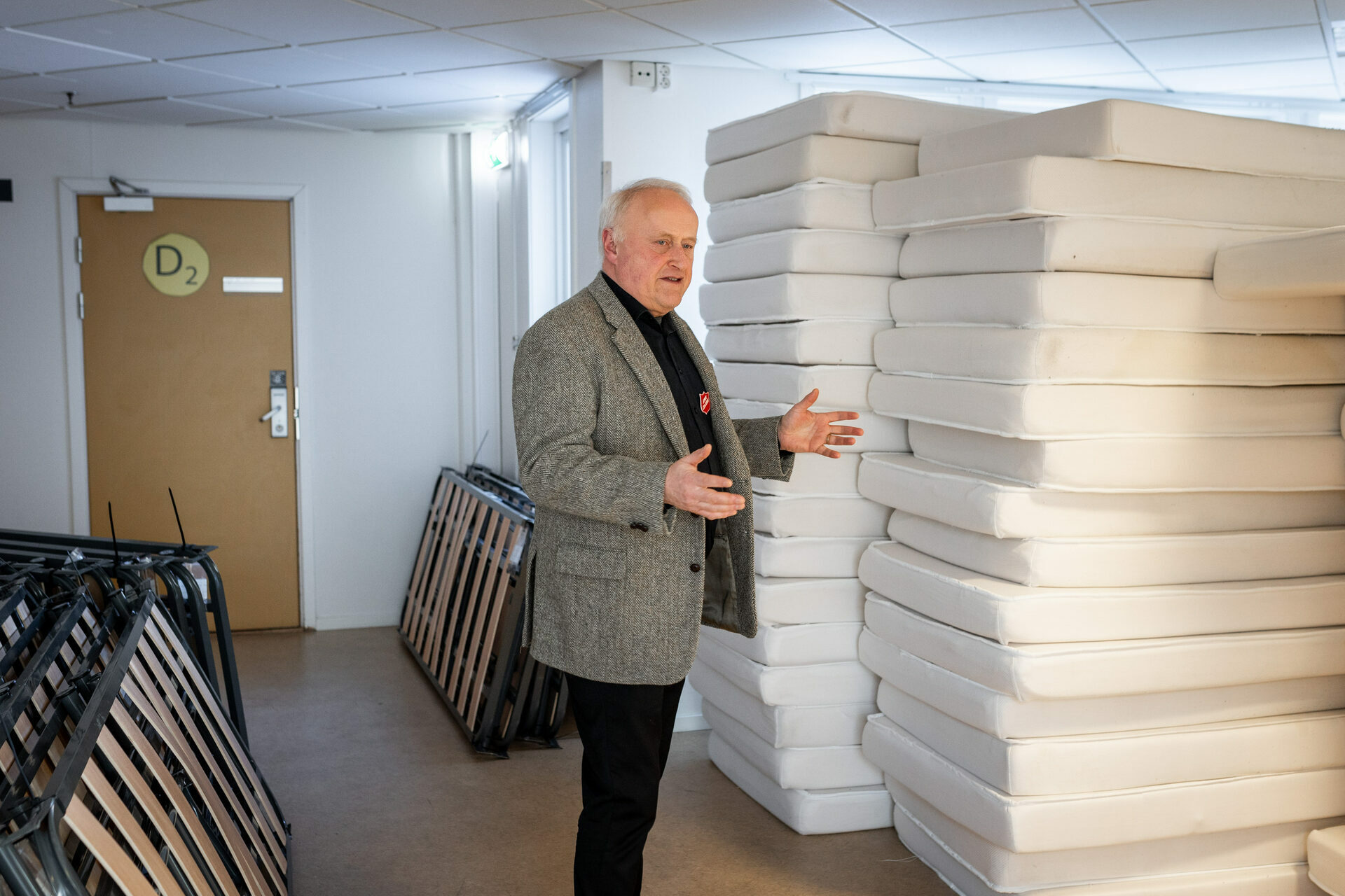 Foto: Mann i 60-årene med grått hår og grønn dressjakke gestikulerer med armene mens han snakker. Han står i et rom med stabler av madrasser og sengebunner.
