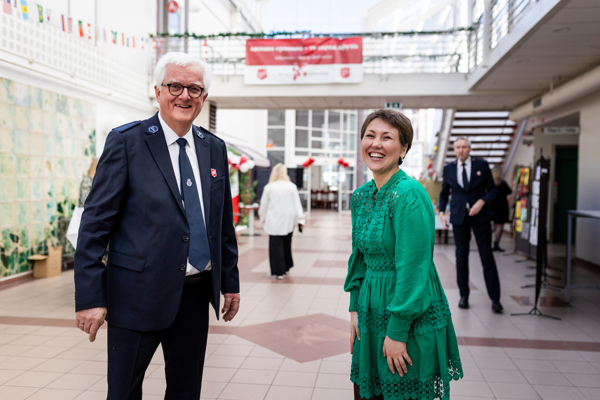 Foto: Eldre mann med grått hår i uniform fra Frelsesarmeen ler sammen med en kvinne i 30-årene. Hun har på seg en grønn kjole, og de står innendørs i en stor hall med glasstak.