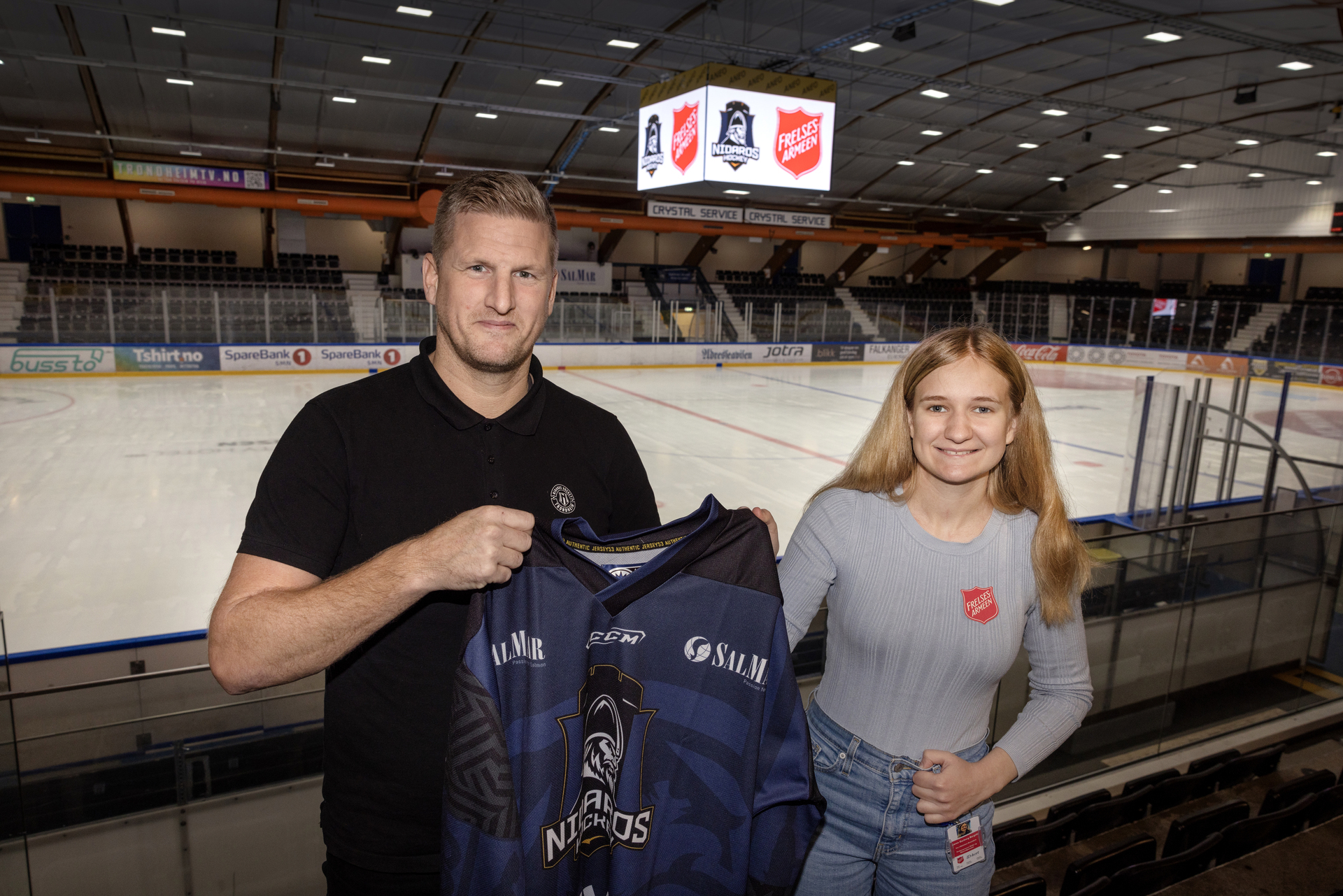 Foto: Samme mann og kvinne står på tribunen med isen i bakgrunnen. I taket vises et skilt med de to logoene på. Han holder opp en blå spillerdrakt og hun har en rød logo fra Frelsesarmeen på brystet.
