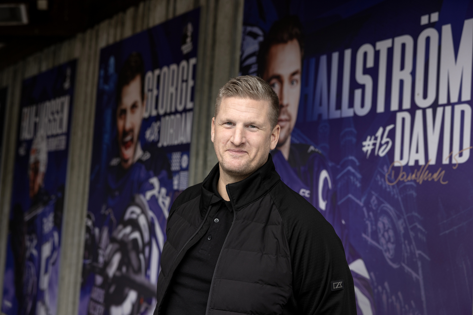 Foto: Mann med blondt hår i 40-årene står i svart skjorte foran en betongvegg med plakater av ishockeystjerner i hver sin blå drakt.