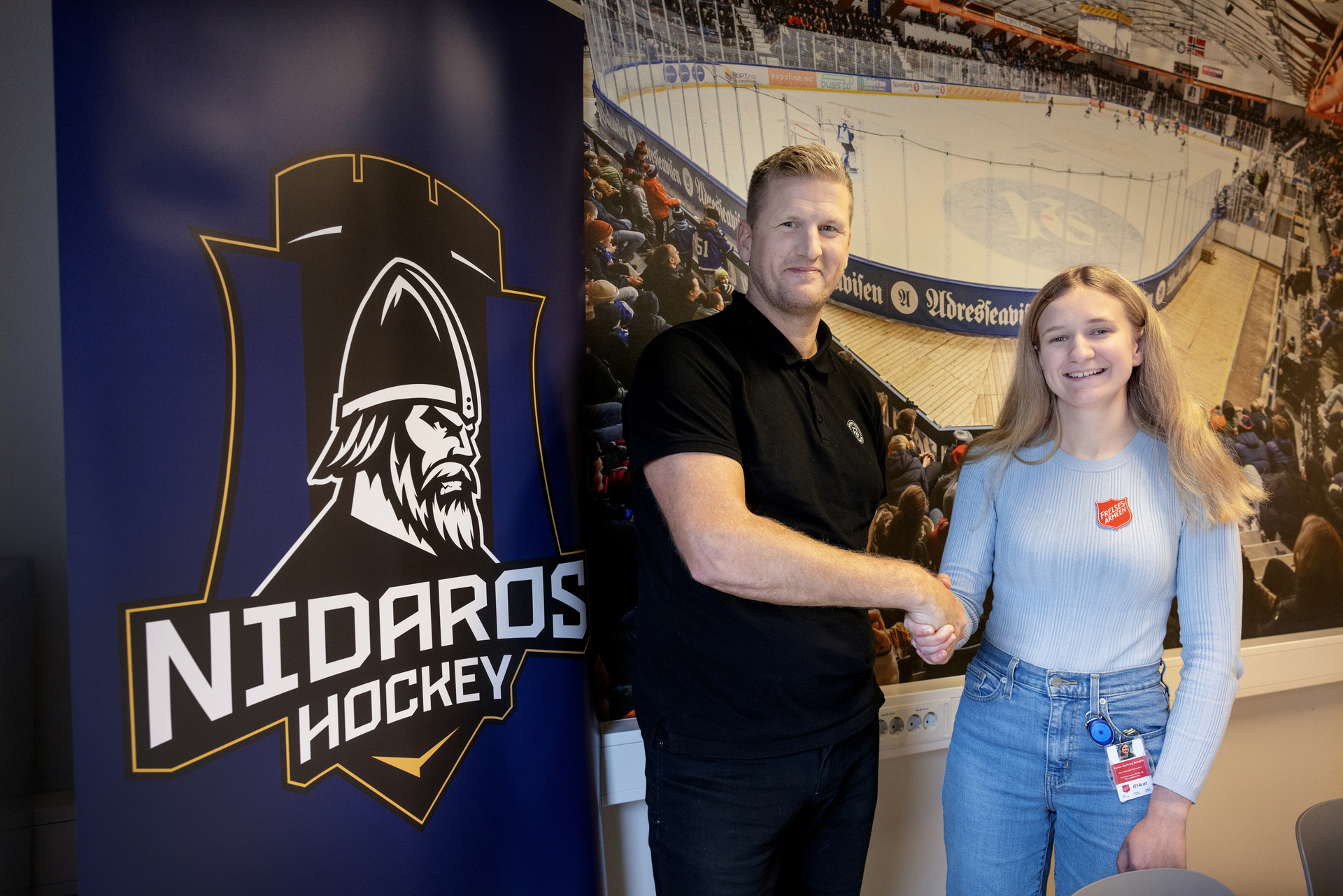 Foto: Blond mann i 40-årene gir blond kvinne i 30-årene et godt håndtrykk. De står foran et bilde av en ishockeybane og en plakat av Nodaros Hockey sin logo av en viking.