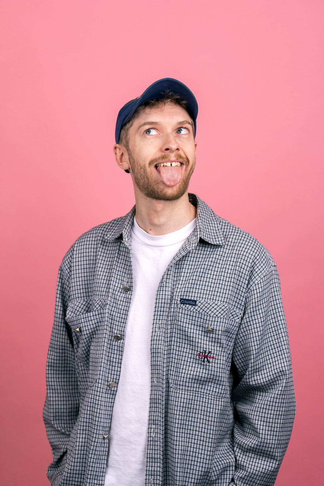 Foto: Mann skjegg, caps og rutete skjorte strekker tunge og ser til siden. Veggen bak ham er rosa.
