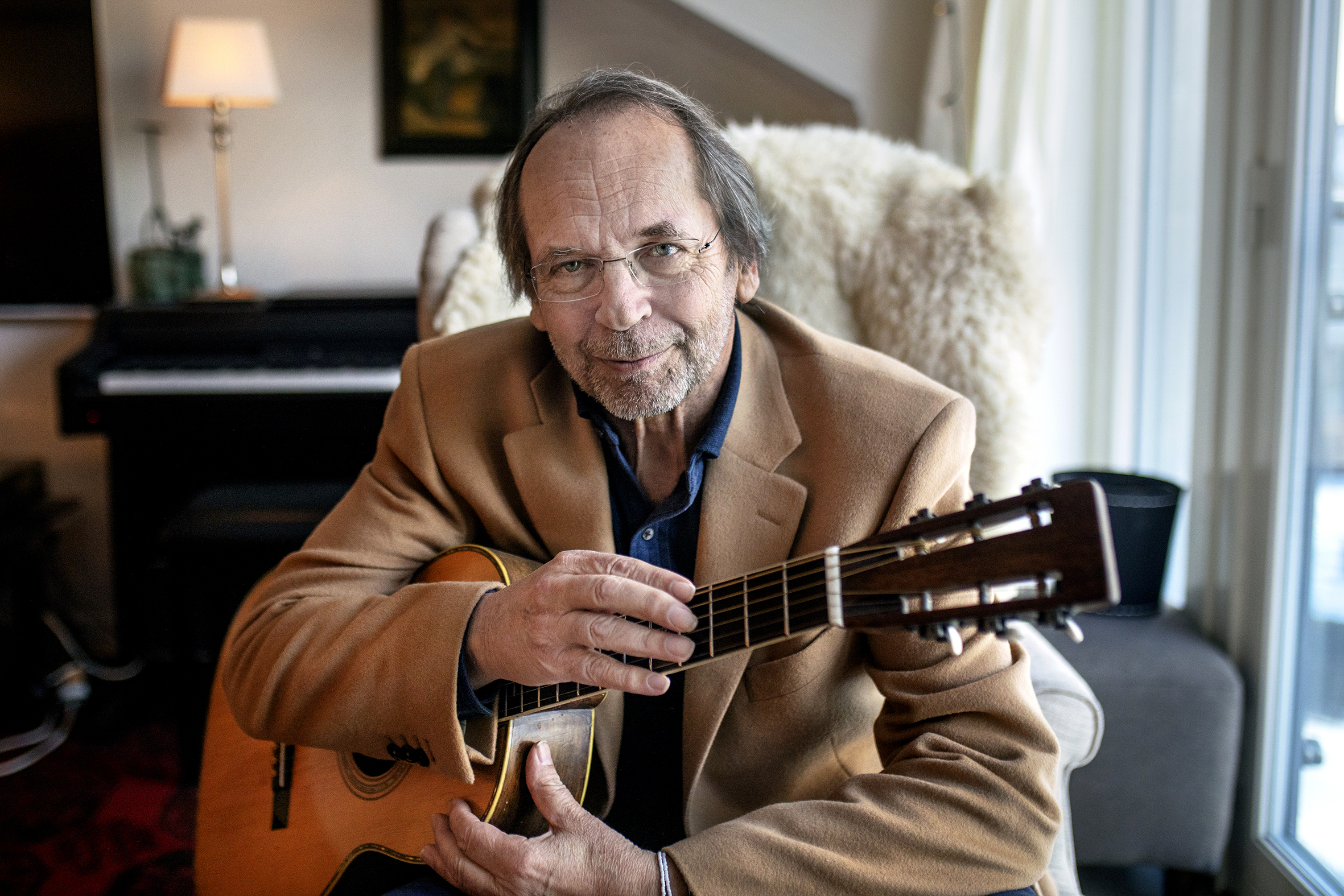 Foto: Mann i 70-årene med tynt, halvlangt hår, briller og en gitar han holder rundt. Han har på seg en beige dressjakke og sitter i en godstol med saueskinn på.