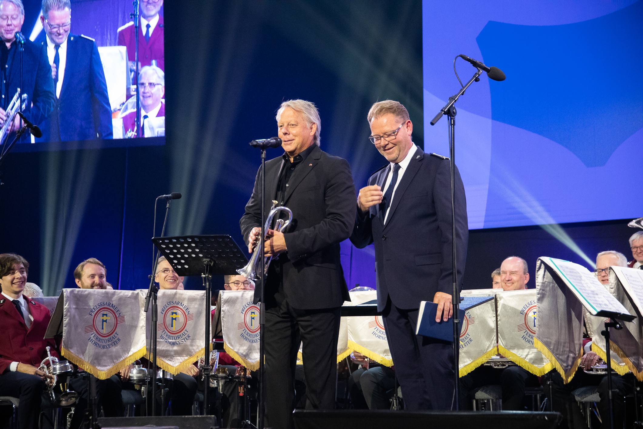 To menn i mørk dress oppe på en scene. I bakgrunnen sitter et orkester fra Frelsesarmeen.