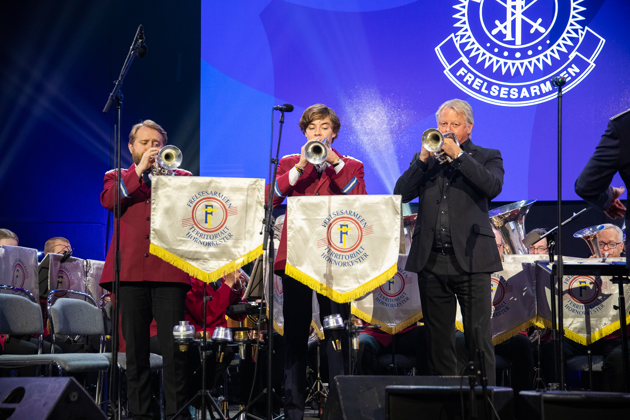 Tre trompetister står på reke foran orkesteret og spiller.