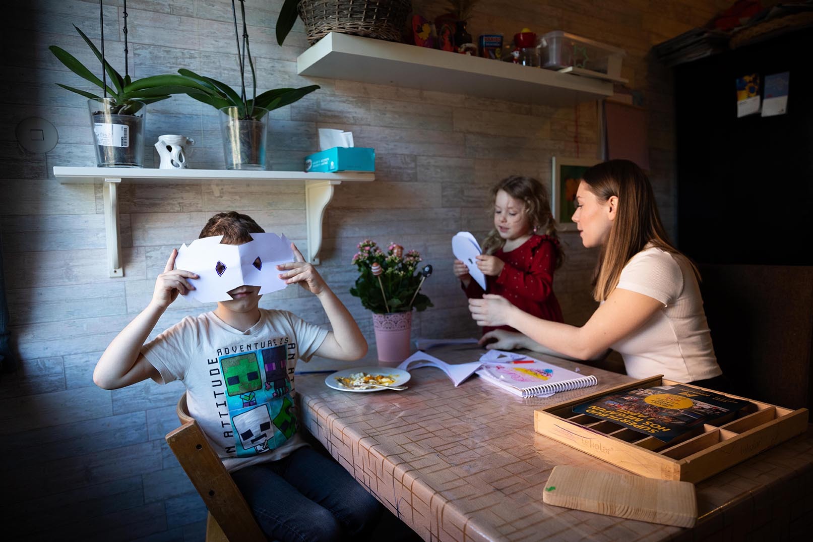 Ei ung jente hjelper ei lita jente med å klippe en papirmaske. En liten gutt tester en ansiktsmaske som er klippet i papir.