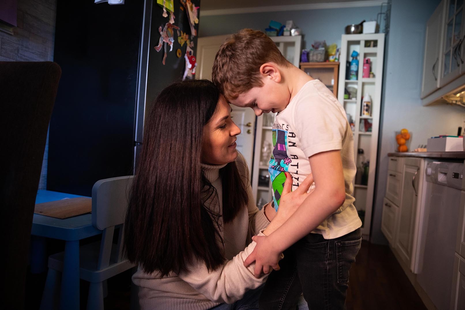 Ung kvinne med mørkt hår har bøyd seg ned på kne. Hun holder en gutt rundt livet og de støtter hodene sammen.