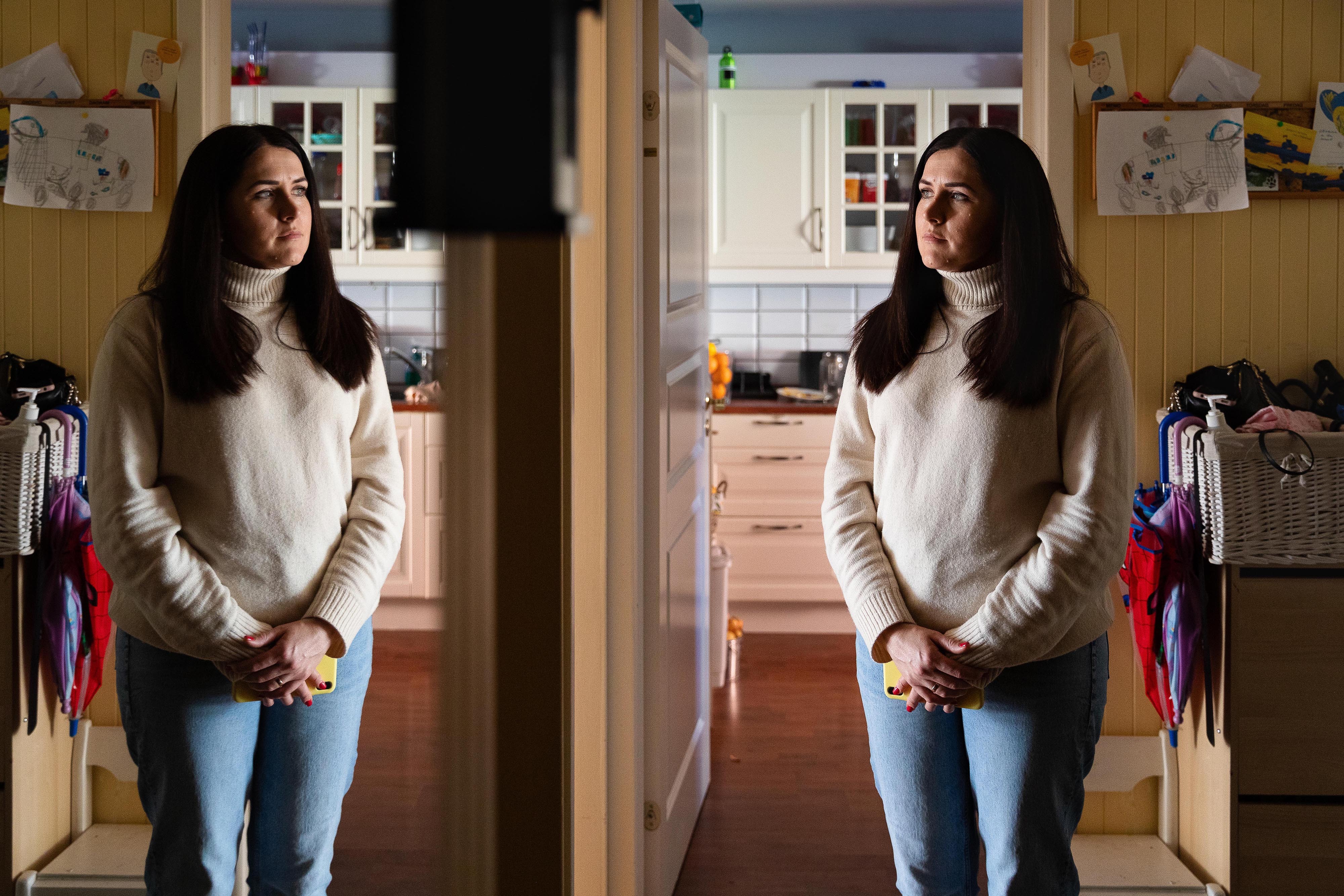 Ung kvinne med mørt hår og høyhalset, hvit genser og dongeribukse står foran et speil i entreen med et kjøkken i bakgrunnen.