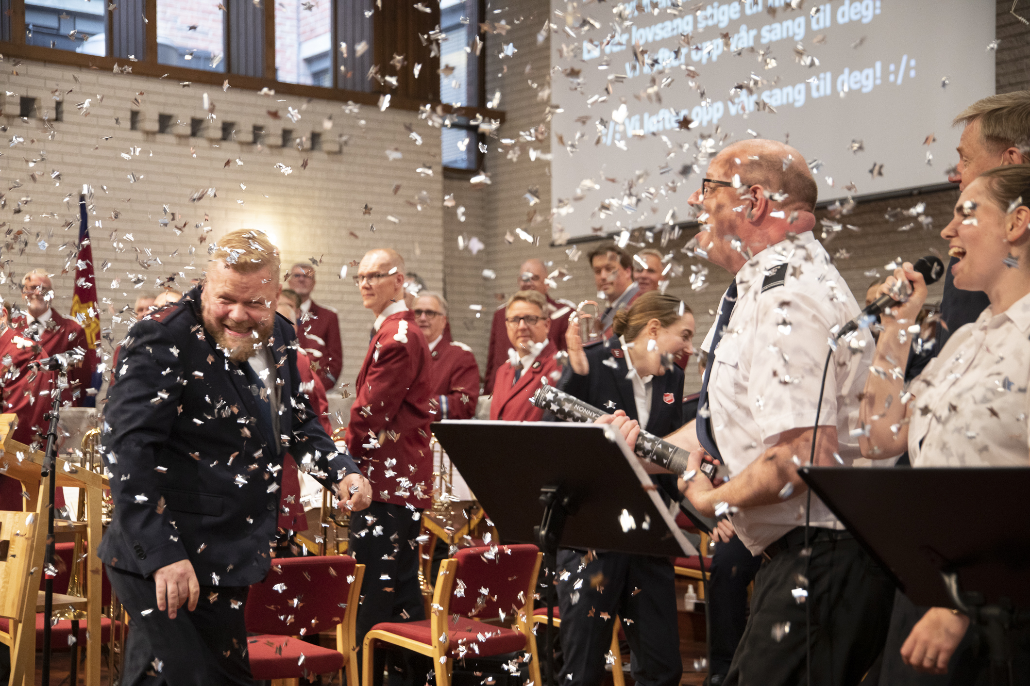 Mann i uniform stråler mot kamera. I luften masse konfetti. Ved siden av et kor som synger.