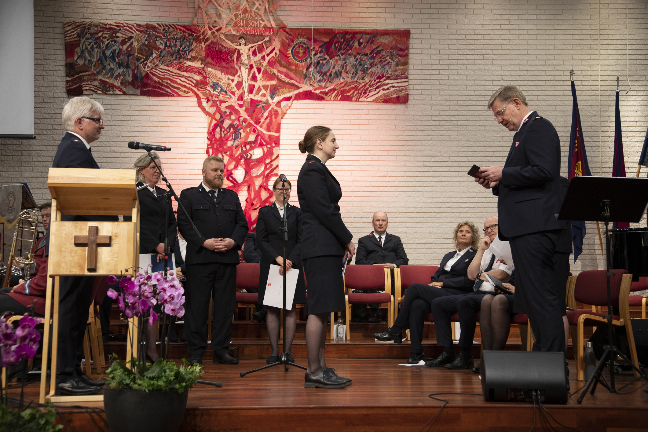 Kirkerom med kors på vegg og talerstol. En kvinne blir ordinert til offiser i Frelsesarmeen. To menn står på hver sin side av henne. Den ene leser fra Bibelen.