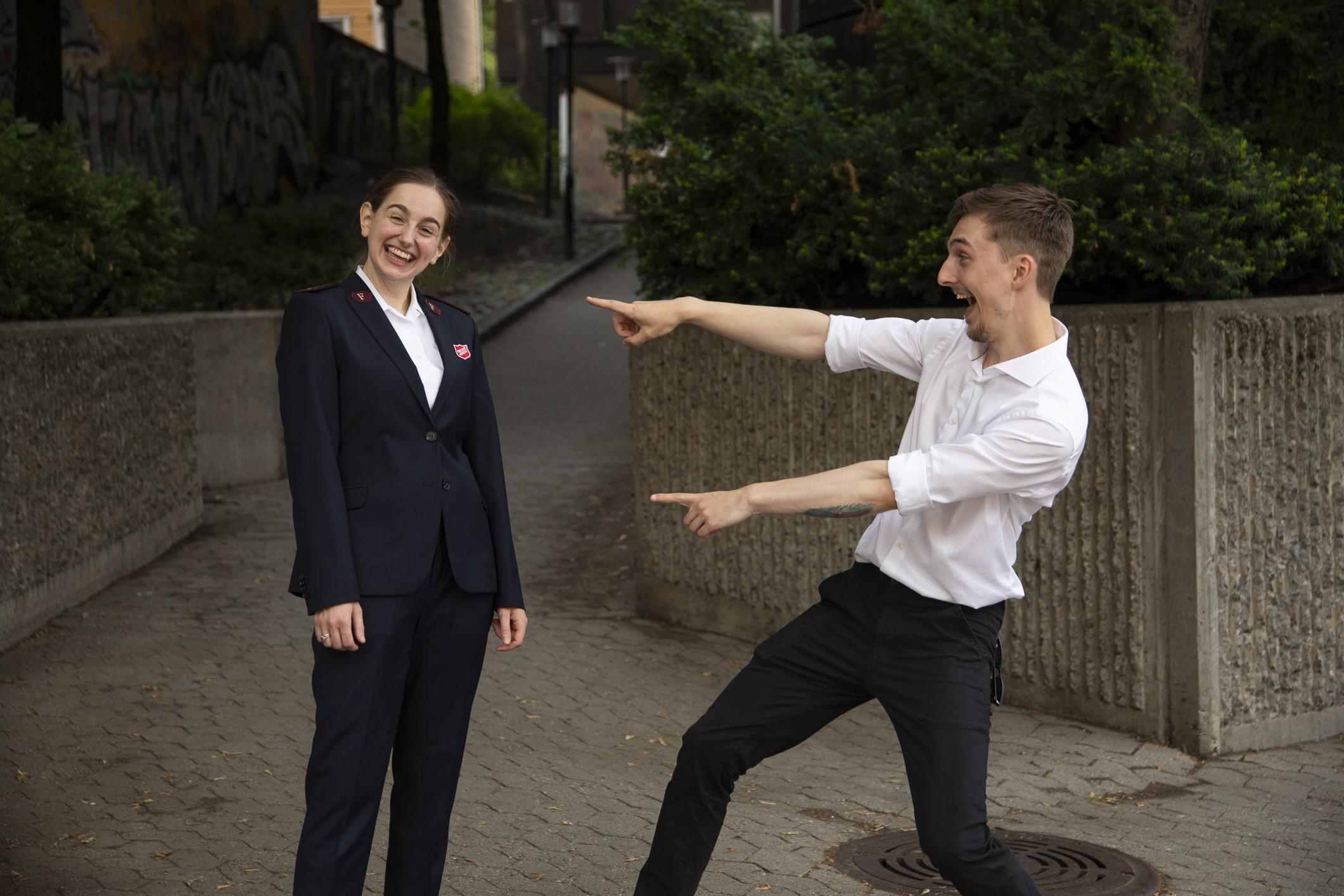 Ung kvinne i uniform oppstilt utendørs foran en passasje. En ung mann i hvit skjorte peker smilende mot henne med begge hender.