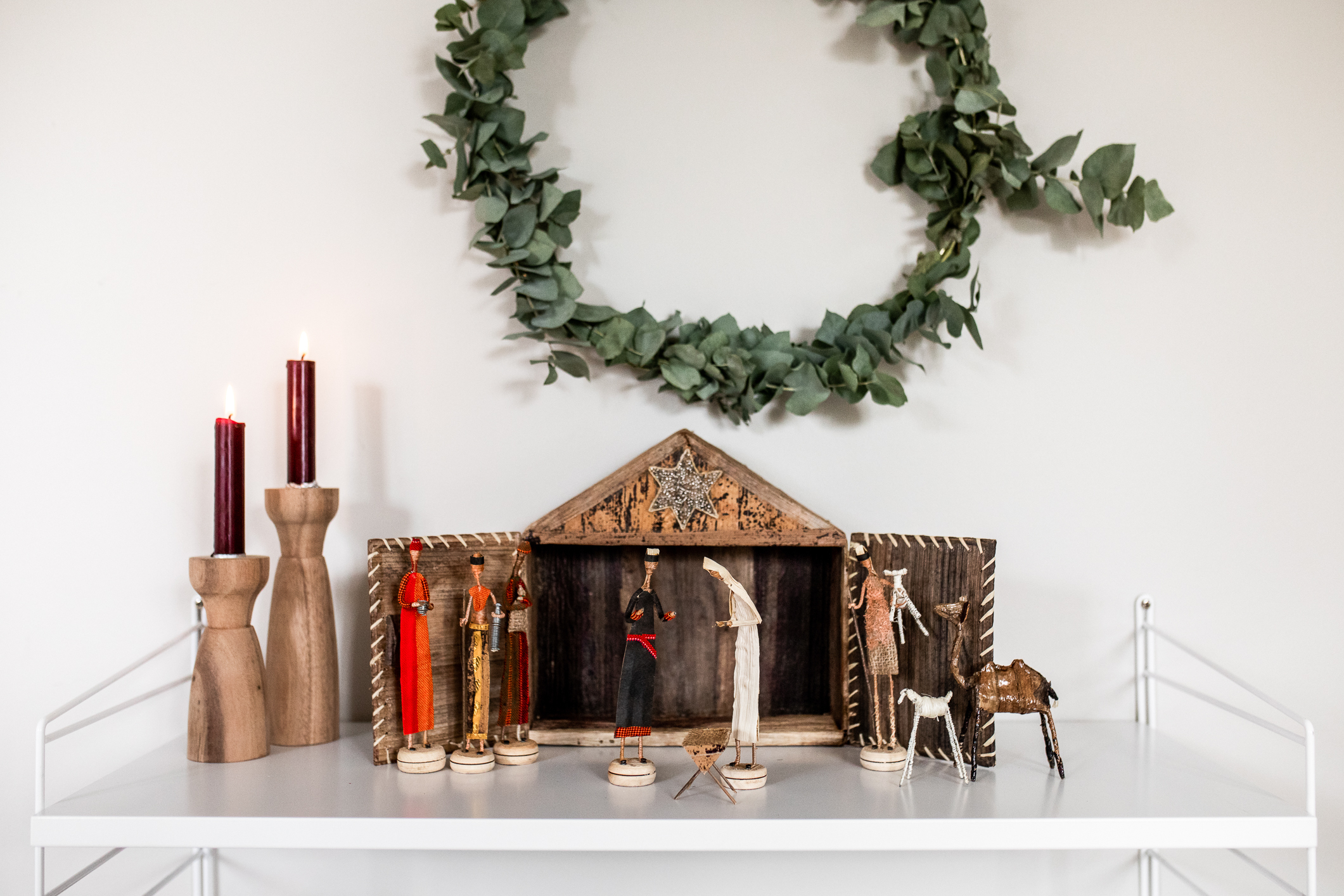 Julekrybbe står oppstilt på en hylle inntil en hvit vegg. Ved siden av står to lysestaker med stearinglys. Over henger en grønn krans av blader.