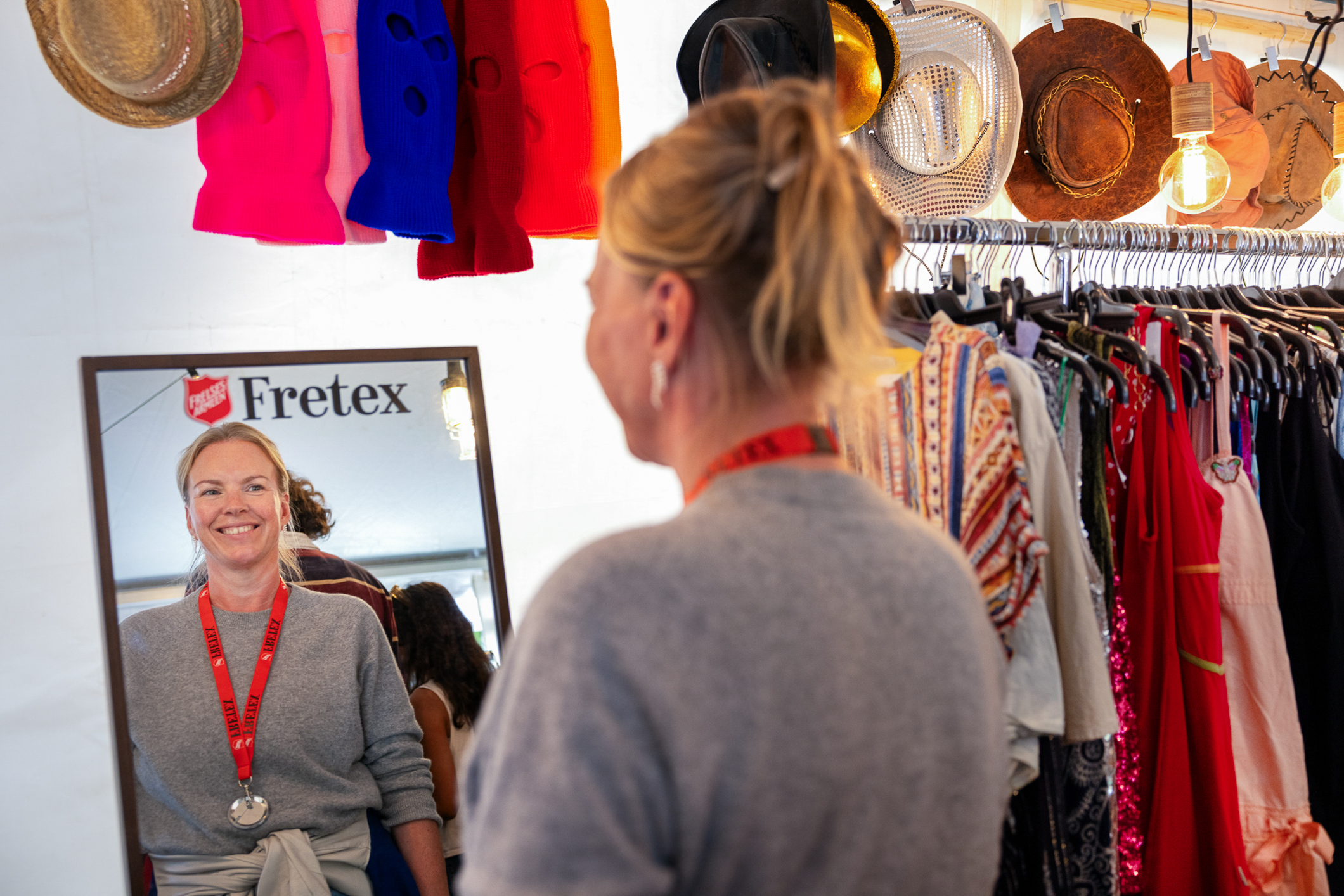 Kvinne som ser seg i speilet. I bakgrunnen henger en rekke hatter og andre plagg.