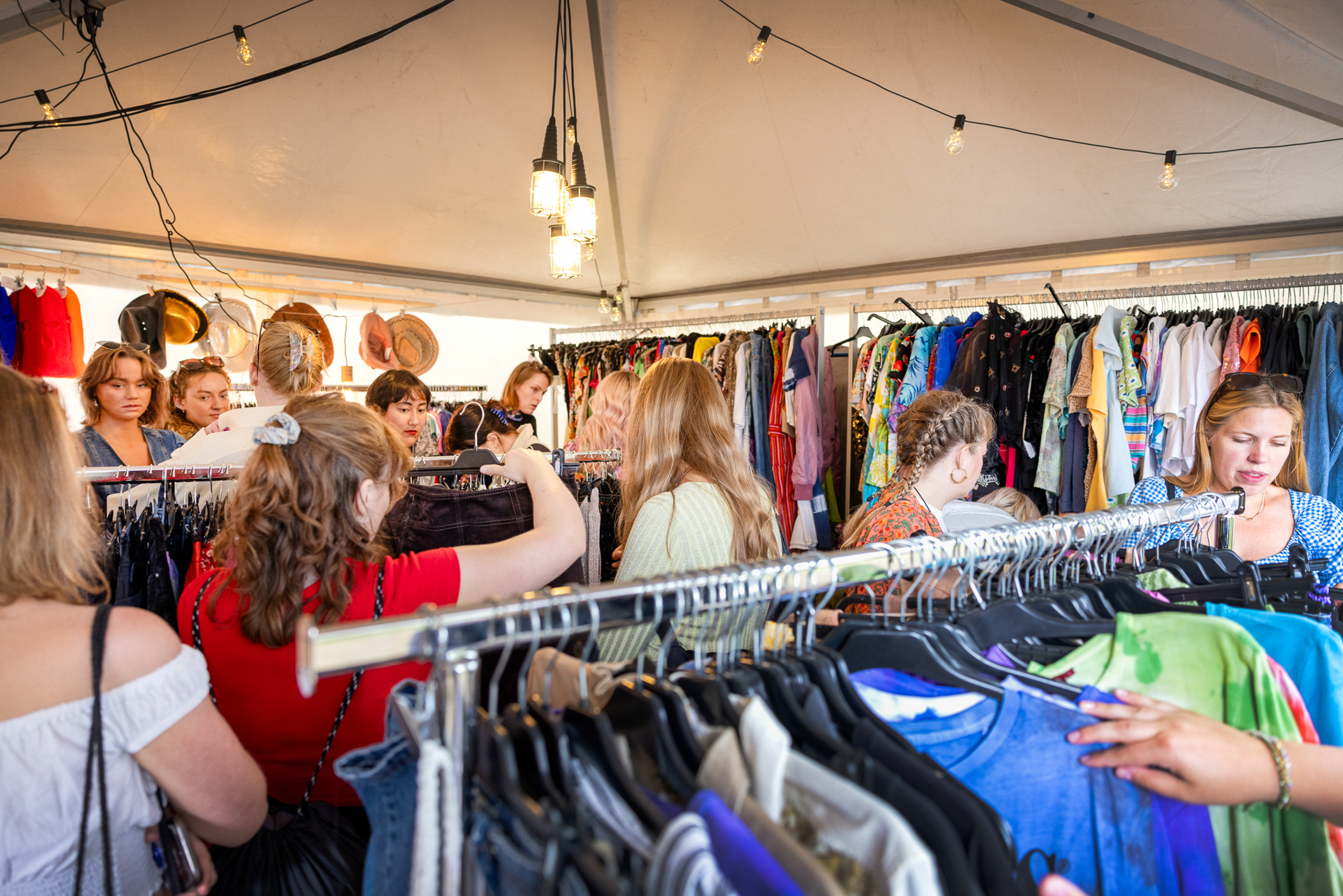 Mange mennesker rundt fargerike klesstativ inni et digert festivaltelt.