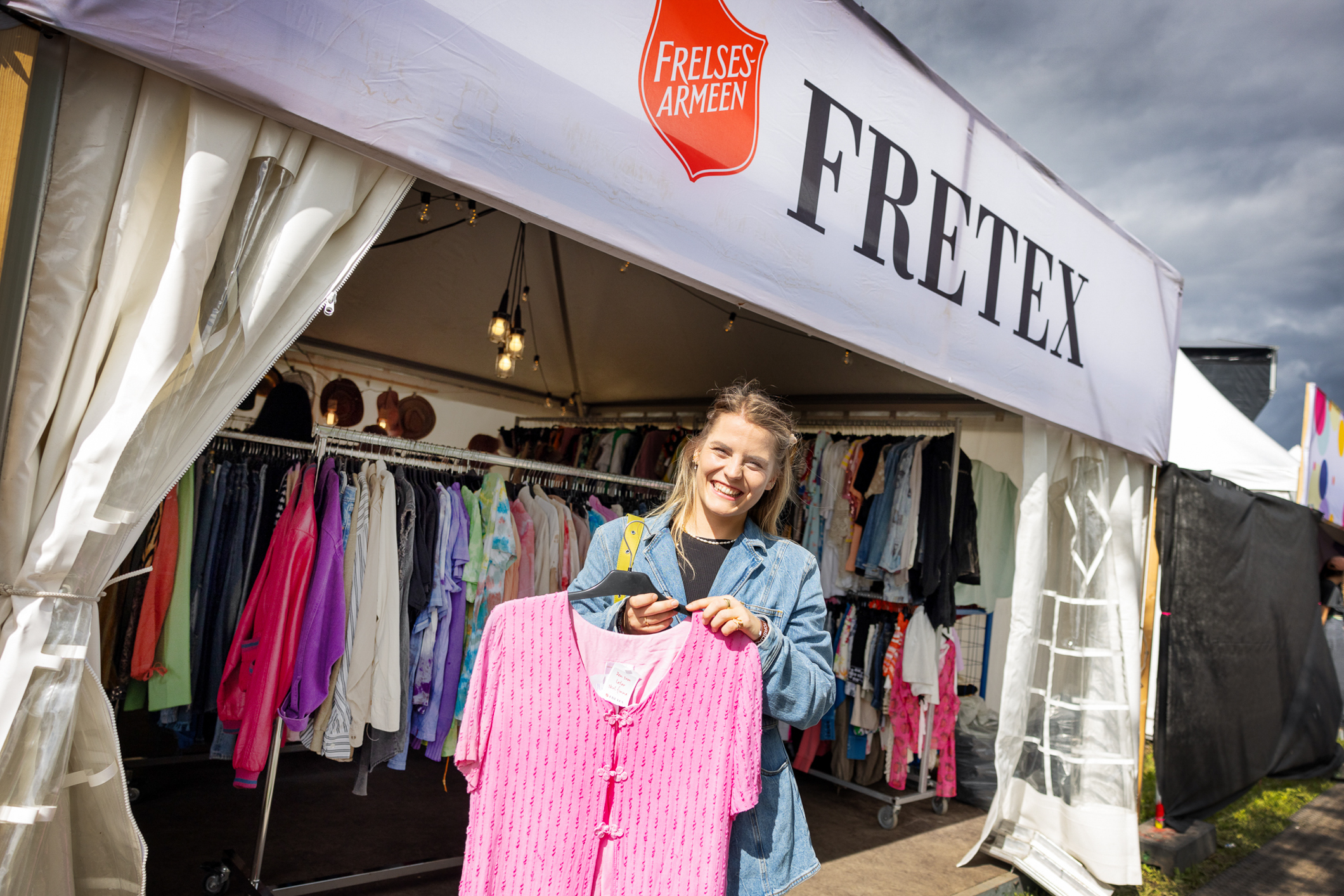 Smilende kvinne utenfor et Fretex-telt på festival. Holder fram en rosa bluse.