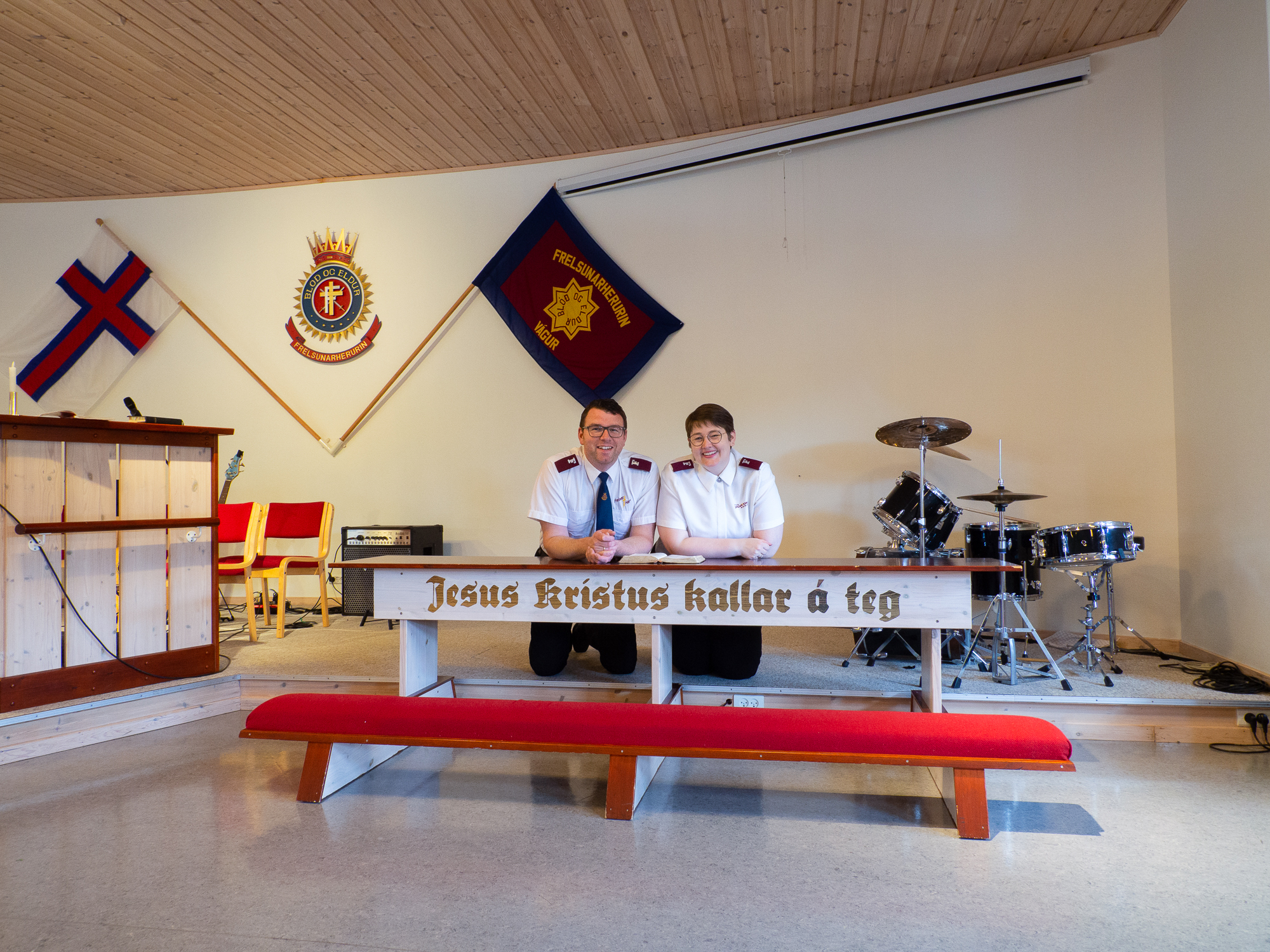 Mann og kvinne kneler på en benk. Foran er gamle, sirlige bokstaver malt med gullfarge.