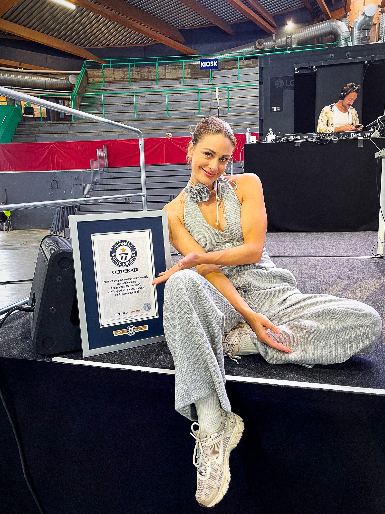 Kvinne som sitter i delvis lotusstilling med buksedress i glitrende sølvstoff. Hun peker på et diplom.