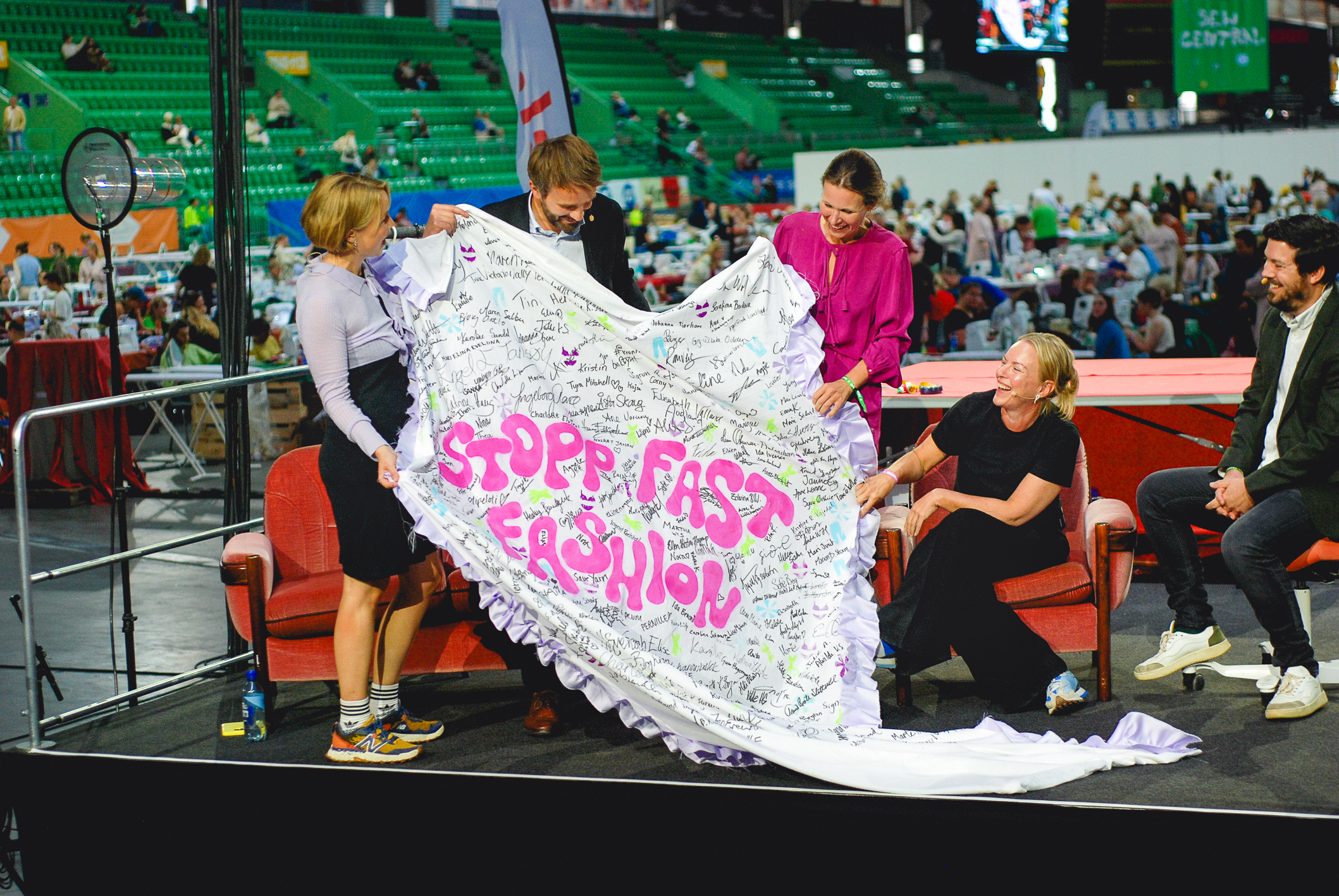 Tre kvinner og en mann holder opp et tøystykke med en rekke underskrifter for å stoppe fast fashion. De sitter på podiet sammen med en mann til.