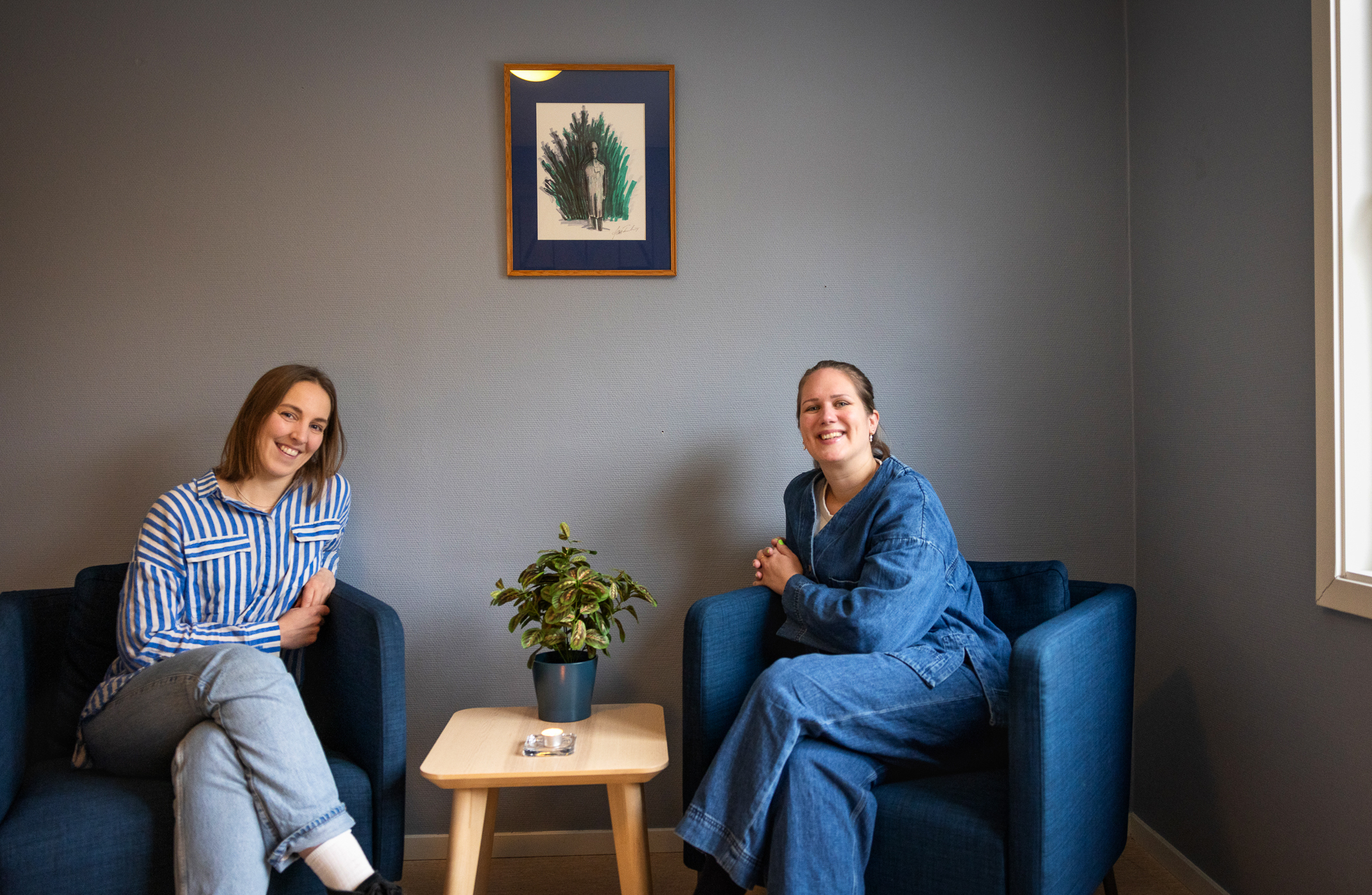 To smilende, unge kvinner sitter i blå lenestoler i et hyggelig rom med et lite kaffebord mellom. Hun til høyre har hestehale og blå dongeriskjorte. Hun til høyre har halvlangt hår og stripete skjorte. Et bilde på veggen.