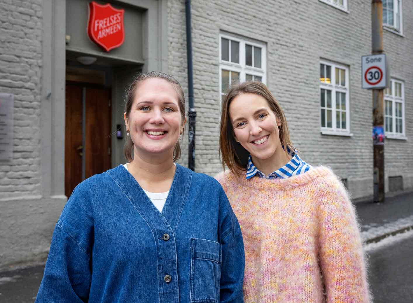 To smilende, unge kvinner utenfor et gråmalt mursteinsbygg som har Frelsesarmeens røde skjold over døren. Hun til venstre har håret bak og blå dongeriskjorte. Hun til høyre har halvlangt hår og ferskenfarget strikkegenser.