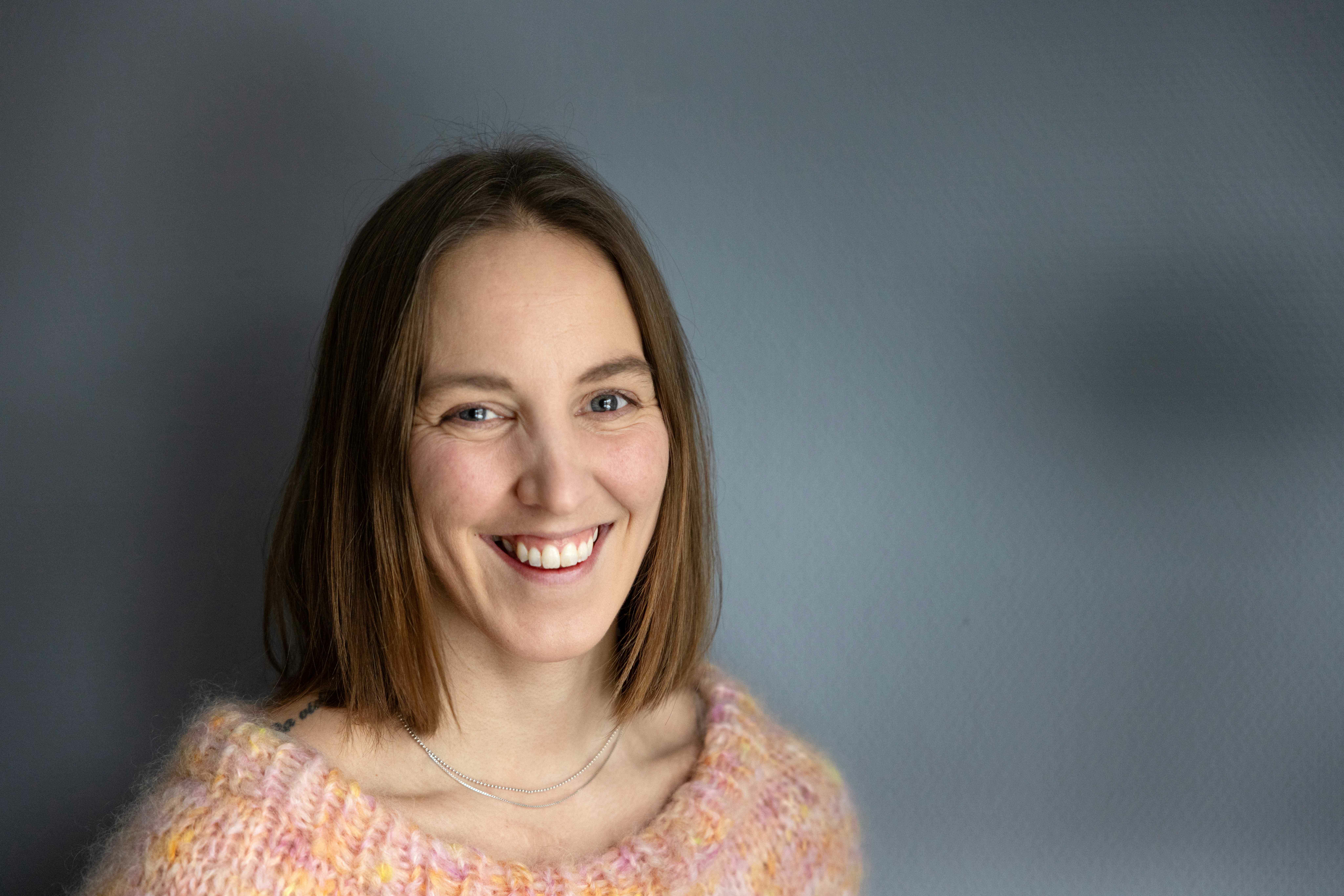 Ung kvinne med halvlangt hår har på seg en ferskenfarget strikkegenser. Veggen bak er grå.