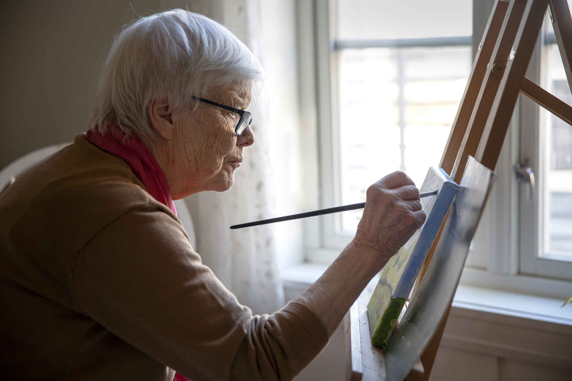 Bildet viser en eldre dame som, i dyp konsentrasjon, sitter fremoverlent og maler på et lerret.