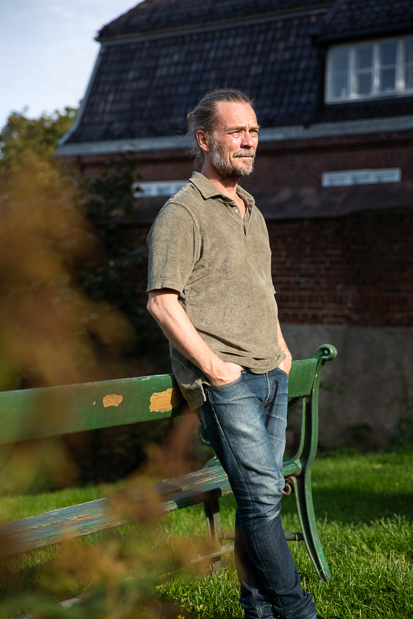 Mann i T-skjorte og dongeribukse står og lener seg mot en gammel hagebenk.