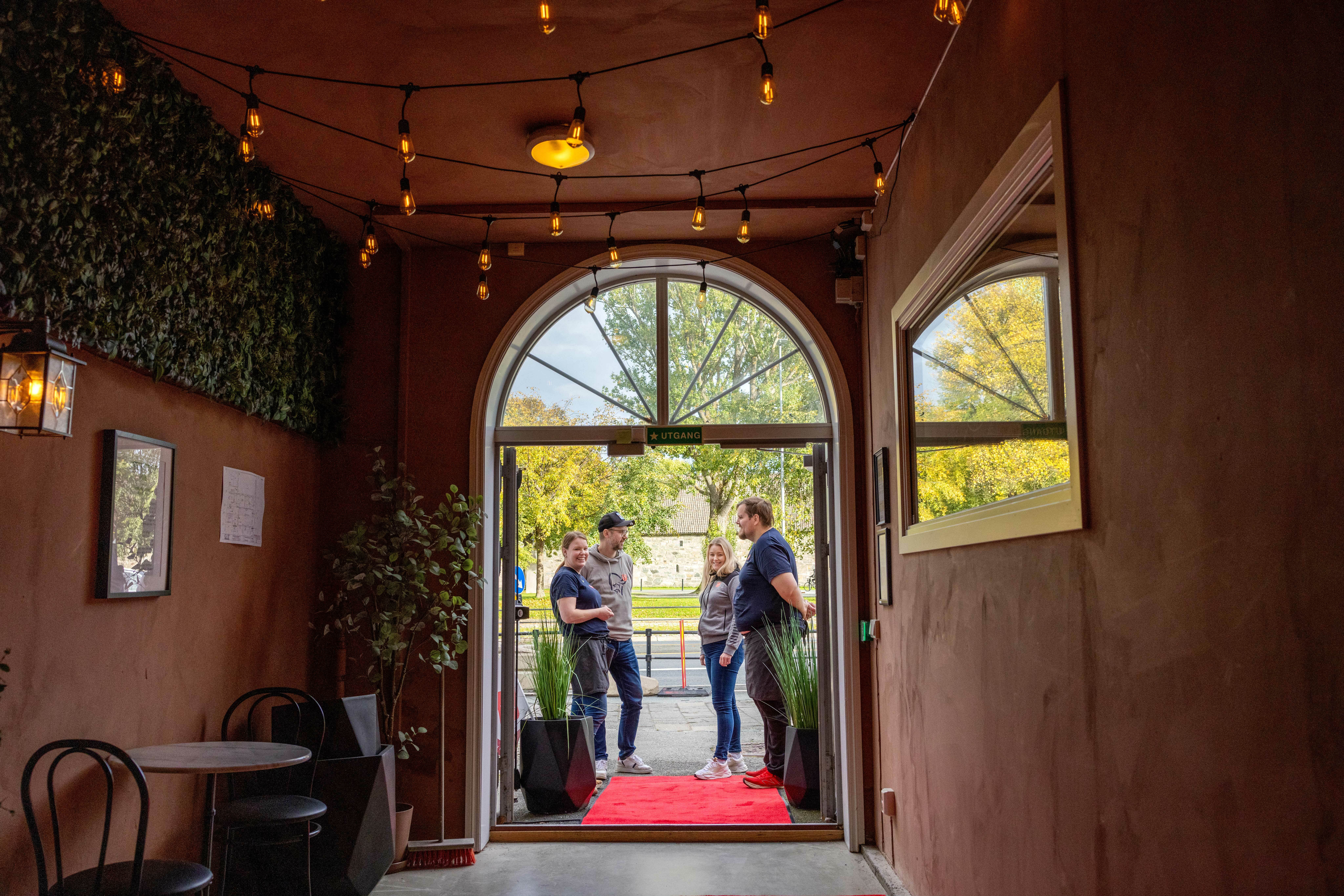 Gjennom en buet dør ser vi fire mennesker står utenfor. Gangen har murvegger og bord og stoler langs siden