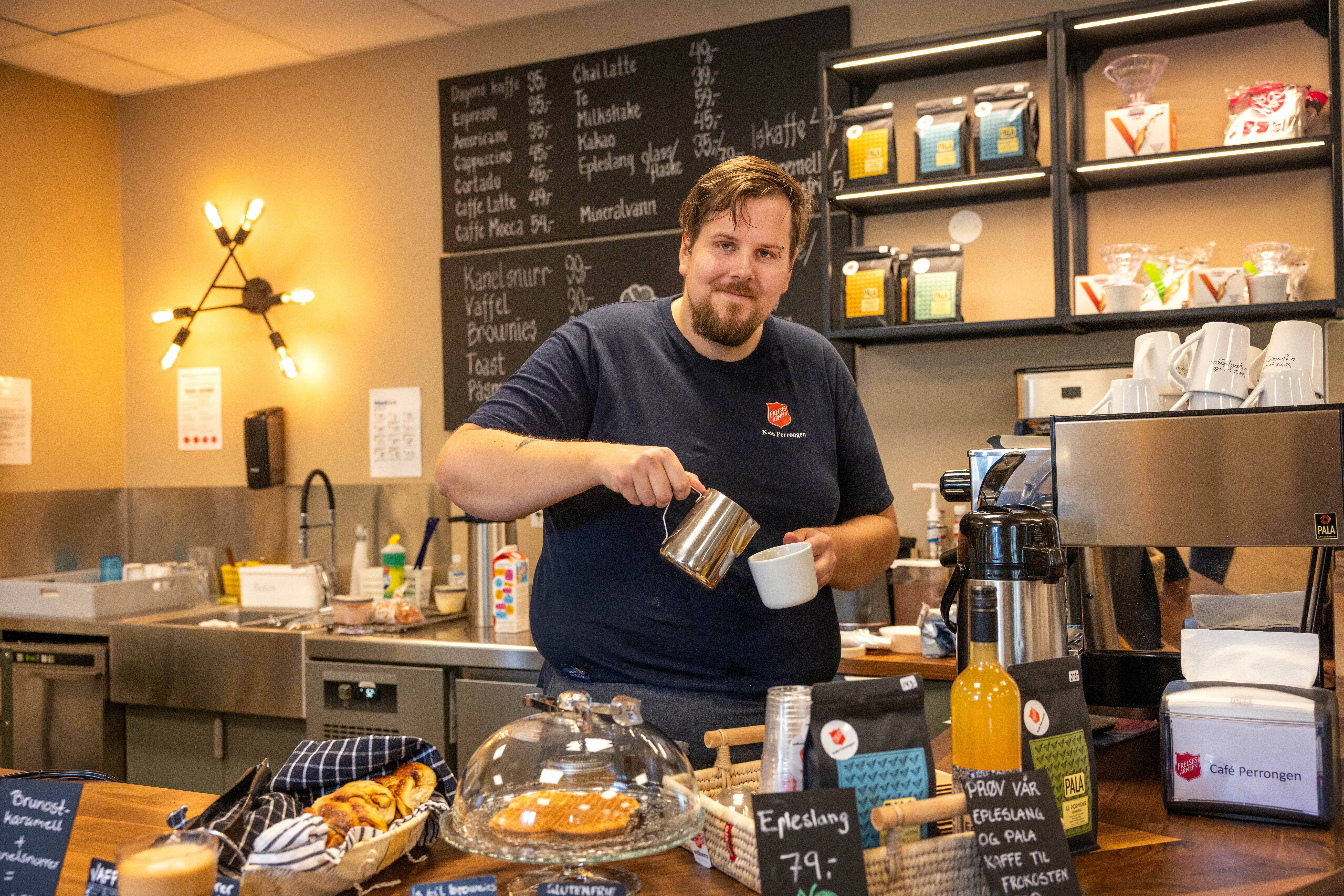 Mann med Frelsesarmeens skjold på brystet står bak disken i en kafé og skjenker kaffe i en kopp.