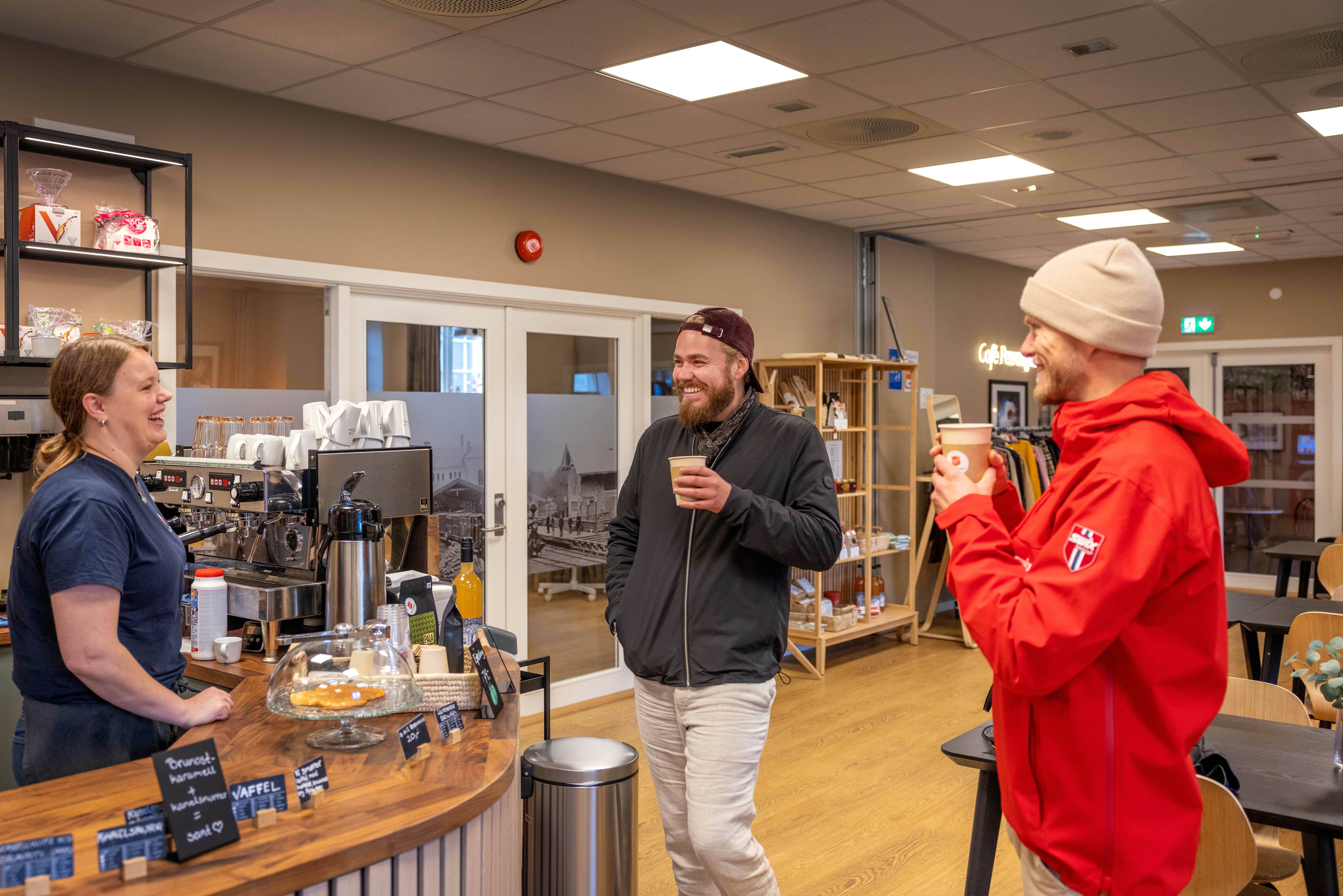 To unge menn drikker kaffe i pappbeger og snakker med en kvinne som står ved disken. Alle smiler.