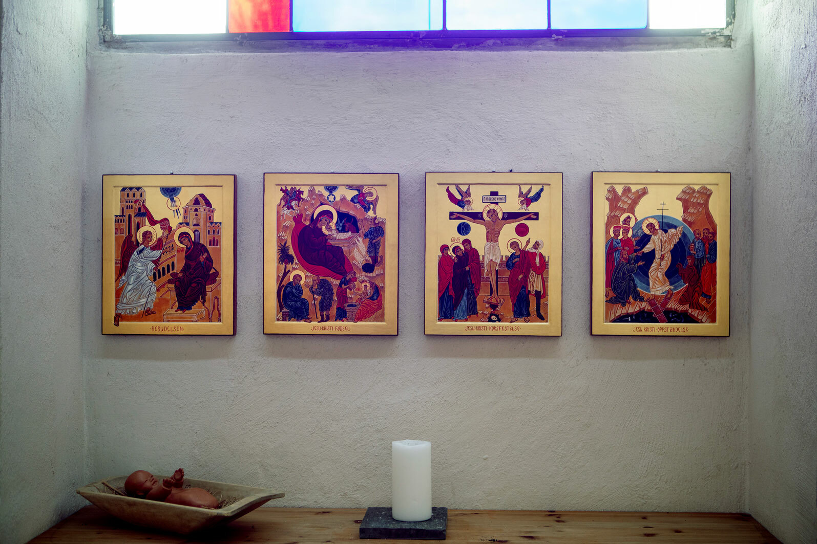 Foto: Fire bilder med religiøse motiv og utstrakt bruk av gullfarge henger på en hvit murvegg. På bordet nedenfor er det et hvitt kubbelys og et fat. Over et vindu med farget glass.