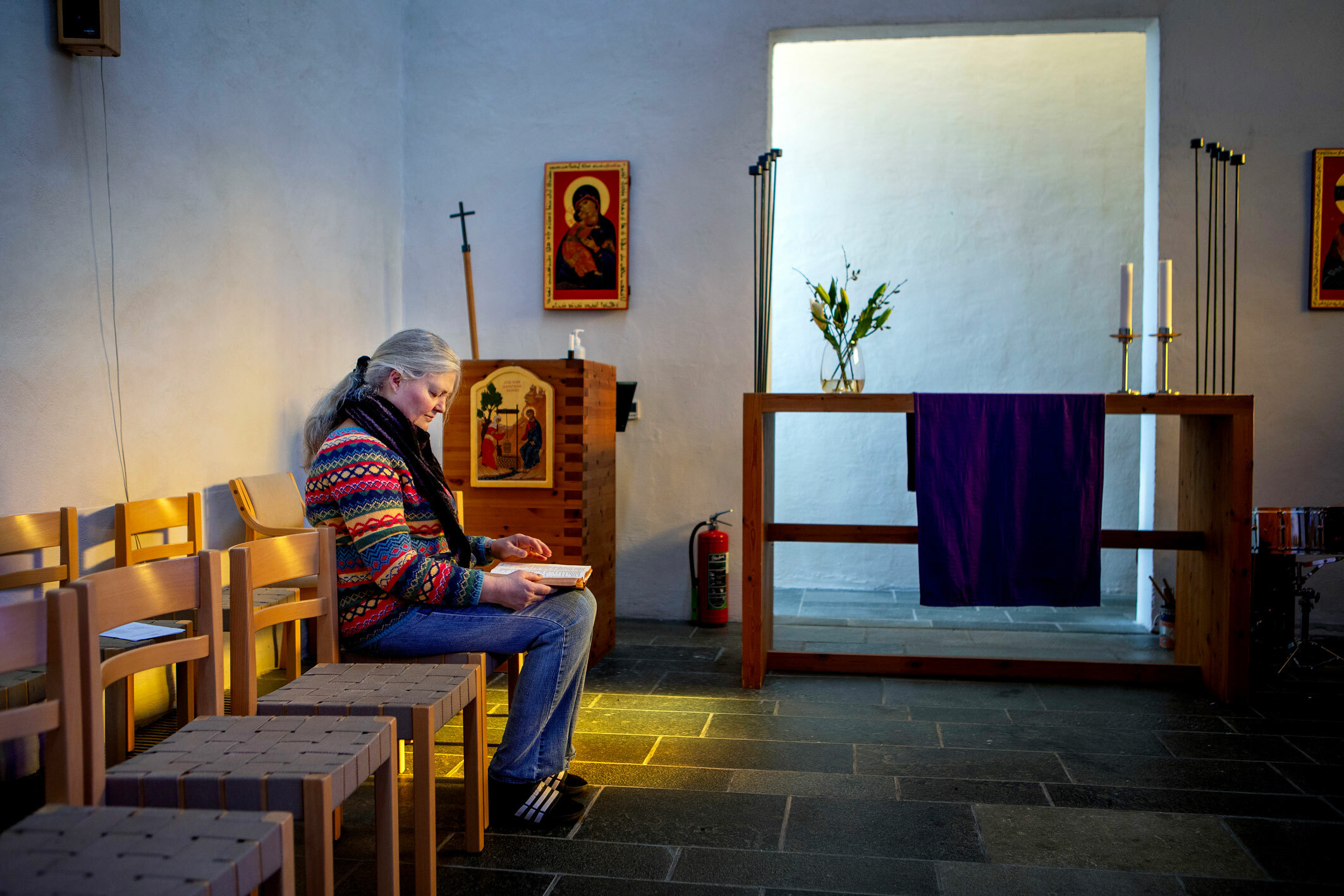 Foto: Kvinne i ullgenser og grått hår i hestehale sitter på en stol inne i den hvitkalkede kapellet. Hun ser ned i en bok hun har på fanget.